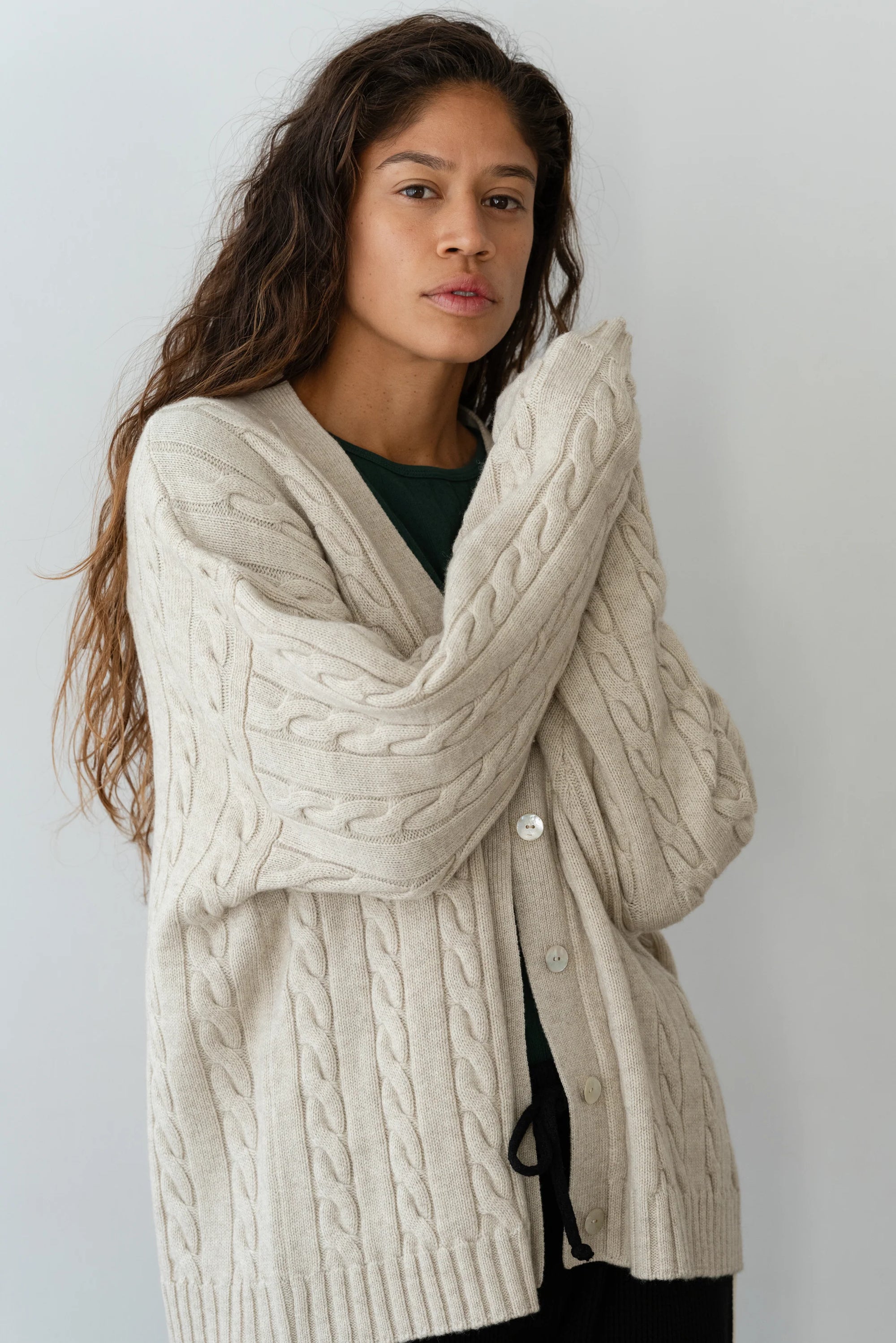 A person with long hair wears The Cable Vneck Card by Donni in beige, showcasing exquisite cable knit detailing over a dark green top. Standing against a light gray background with arms crossed, they maintain a calm expression.