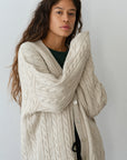 A person with long hair wears The Cable Vneck Card by Donni in beige, showcasing exquisite cable knit detailing over a dark green top. Standing against a light gray background with arms crossed, they maintain a calm expression.