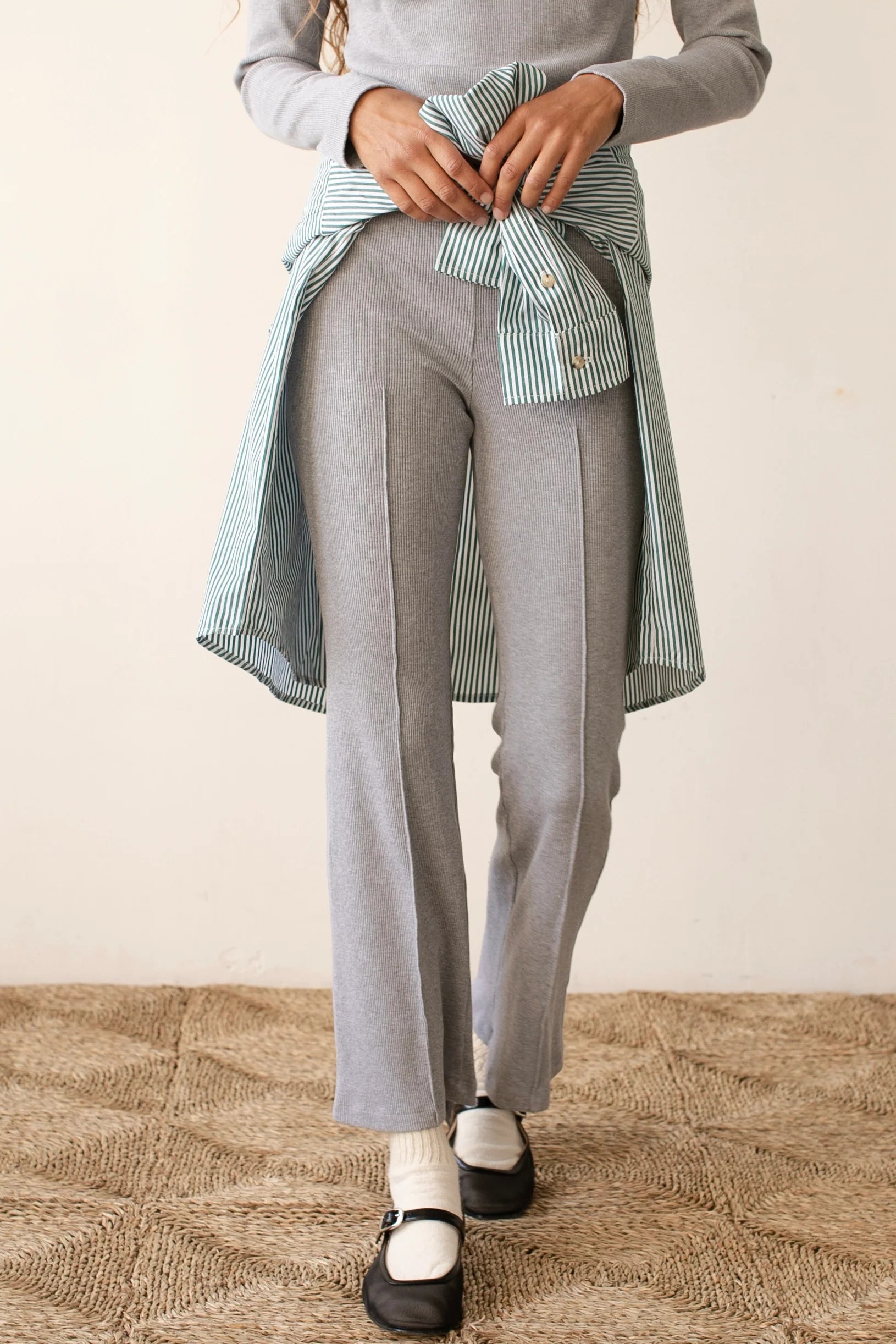 A person wearing Donni&#39;s The Rib Kick Flare in gray pairs it with a light gray top and a striped blue shirt tied around their waist. The look is finished with white socks and black shoes on a textured floor, perfectly blending comfort and style.