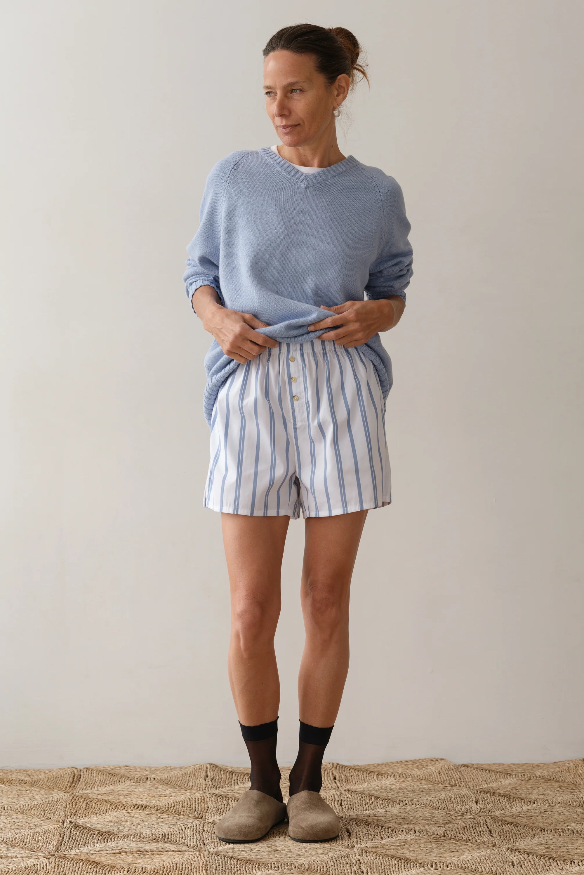 A person stands indoors against a plain background, wearing Donni's light blue sweater paired with The Pop Boxer shorts, crafted in Los Angeles. Their hair is tied back and they wear socks with brown shoes, creating a relaxed and effortlessly composed look.