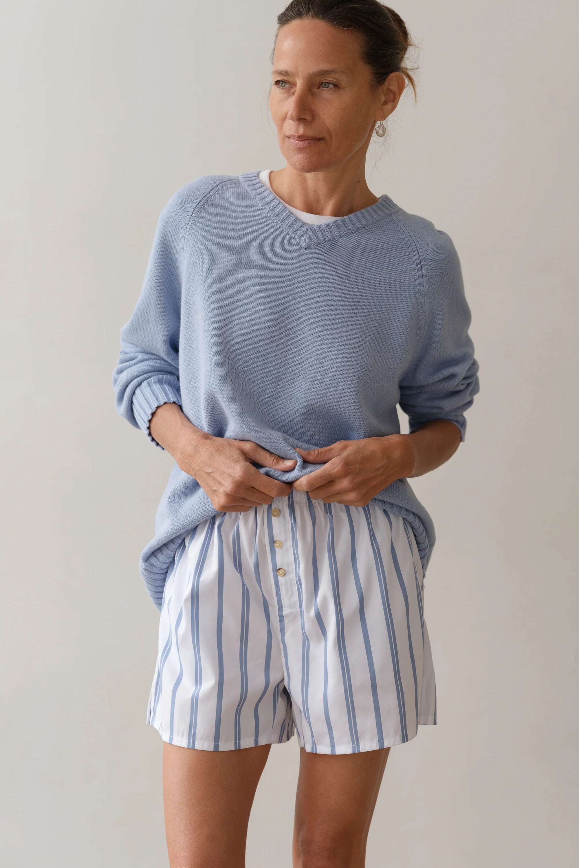 Sporting Donni's The Pop Boxer, a person in a light blue sweater and white cotton poplin shorts with blue stripes poses against a plain backdrop. Made in Los Angeles, the relaxed fit enhances their serene expression as they lightly grasp the waistband of their shorts.