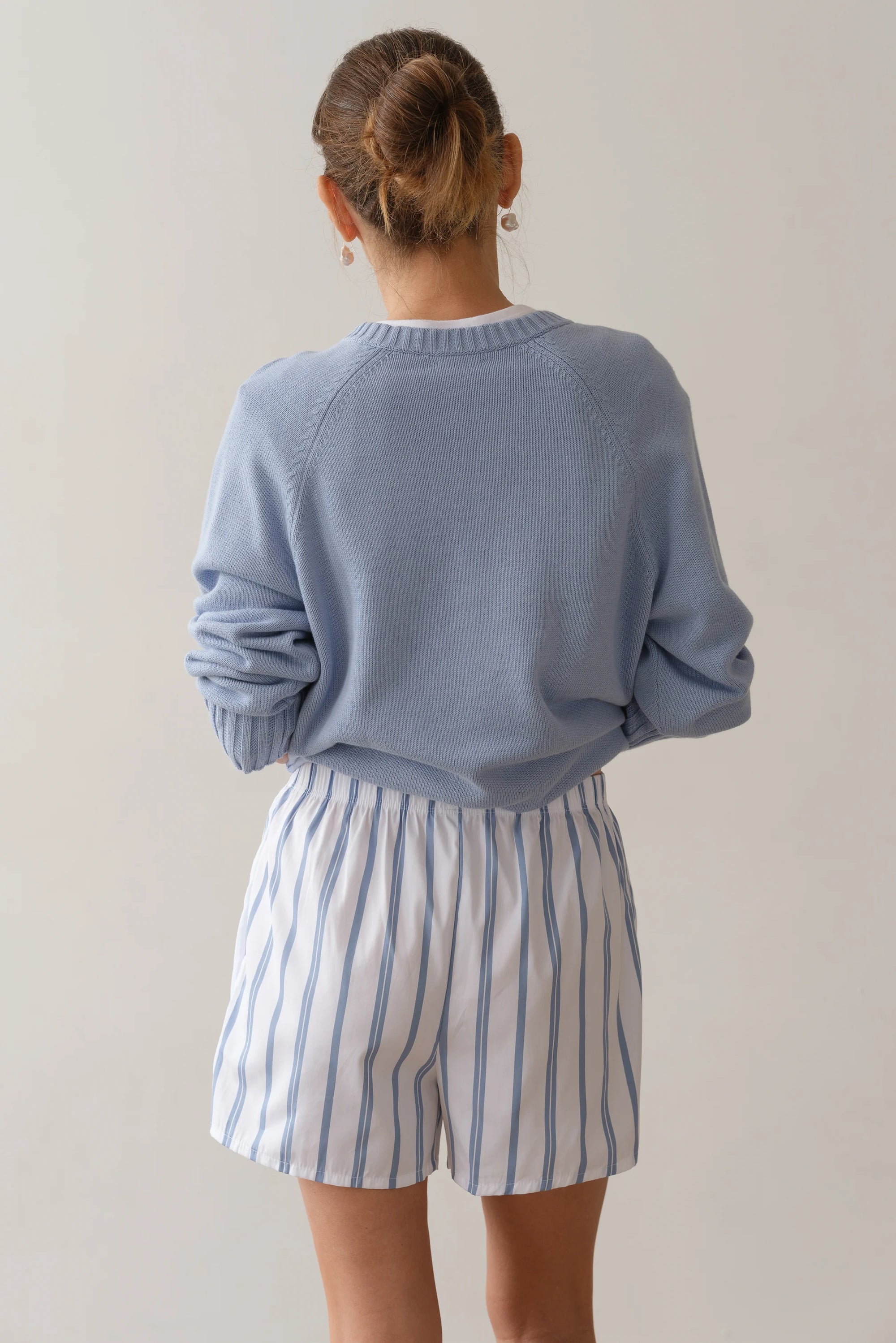 Facing away, a person with a bun hairstyle wears The Pop Boxer by Donni—white shorts with blue vertical stripes—and a light blue cotton poplin sweater. Made in Los Angeles, the ensemble is styled against a neutral background for a simple, minimalist look.