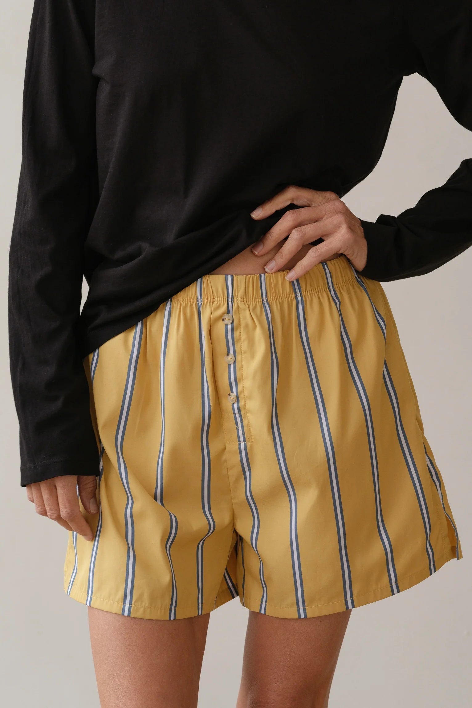 A person in a black long-sleeve shirt holds the waistband of The Pop Boxer by Donni, featuring relaxed-fit yellow shorts with blue and white stripes. Made from breathable cotton poplin, these shorts contrast with the plain, neutral background.