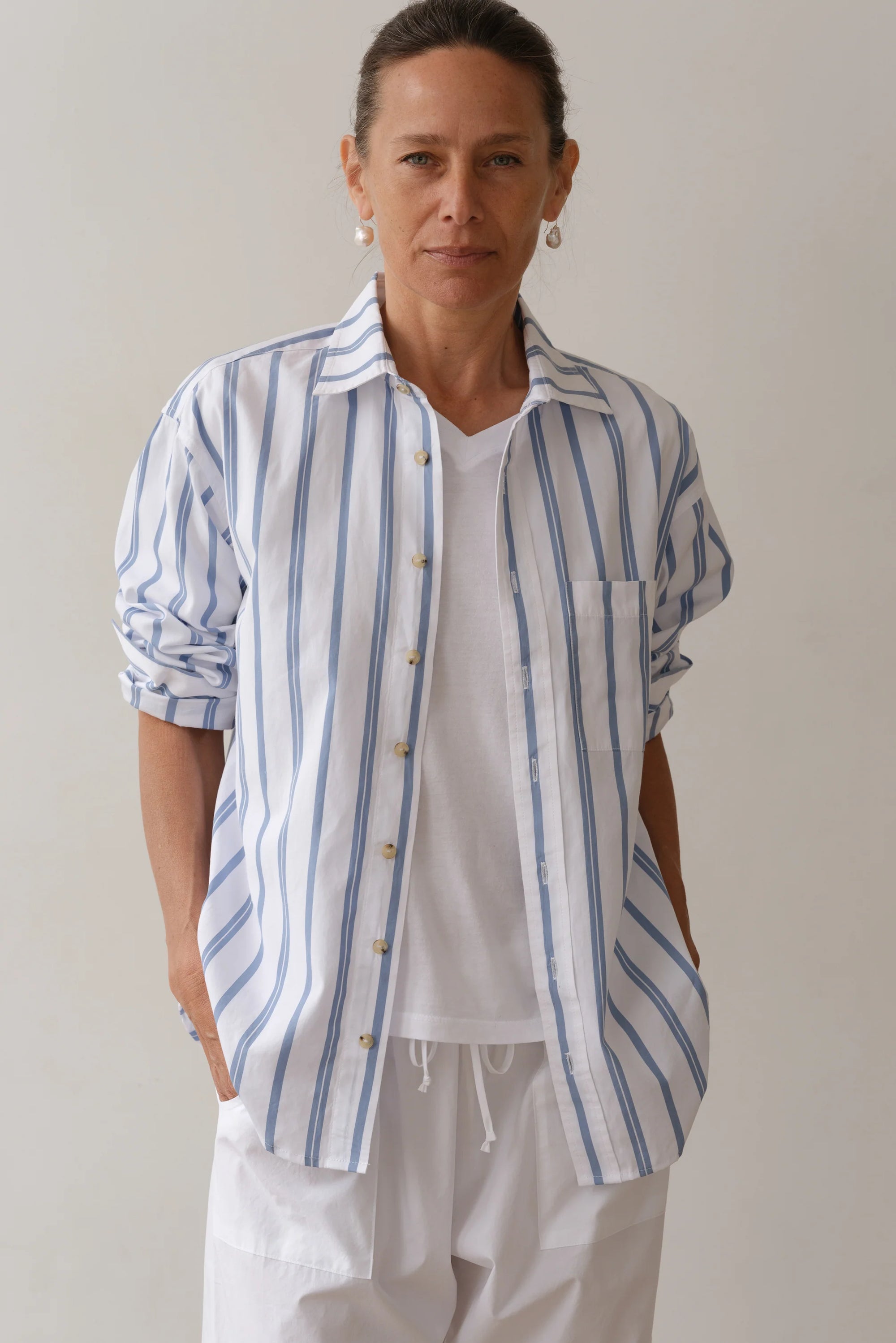 In a relaxed pose, the individual wears Donni's The Poplin Shirt, a blue and white striped button-up made from vintage men's shirting, over a white tee and matching pants. Earrings peek out from their pulled-back hair against a plain, neutral backdrop.