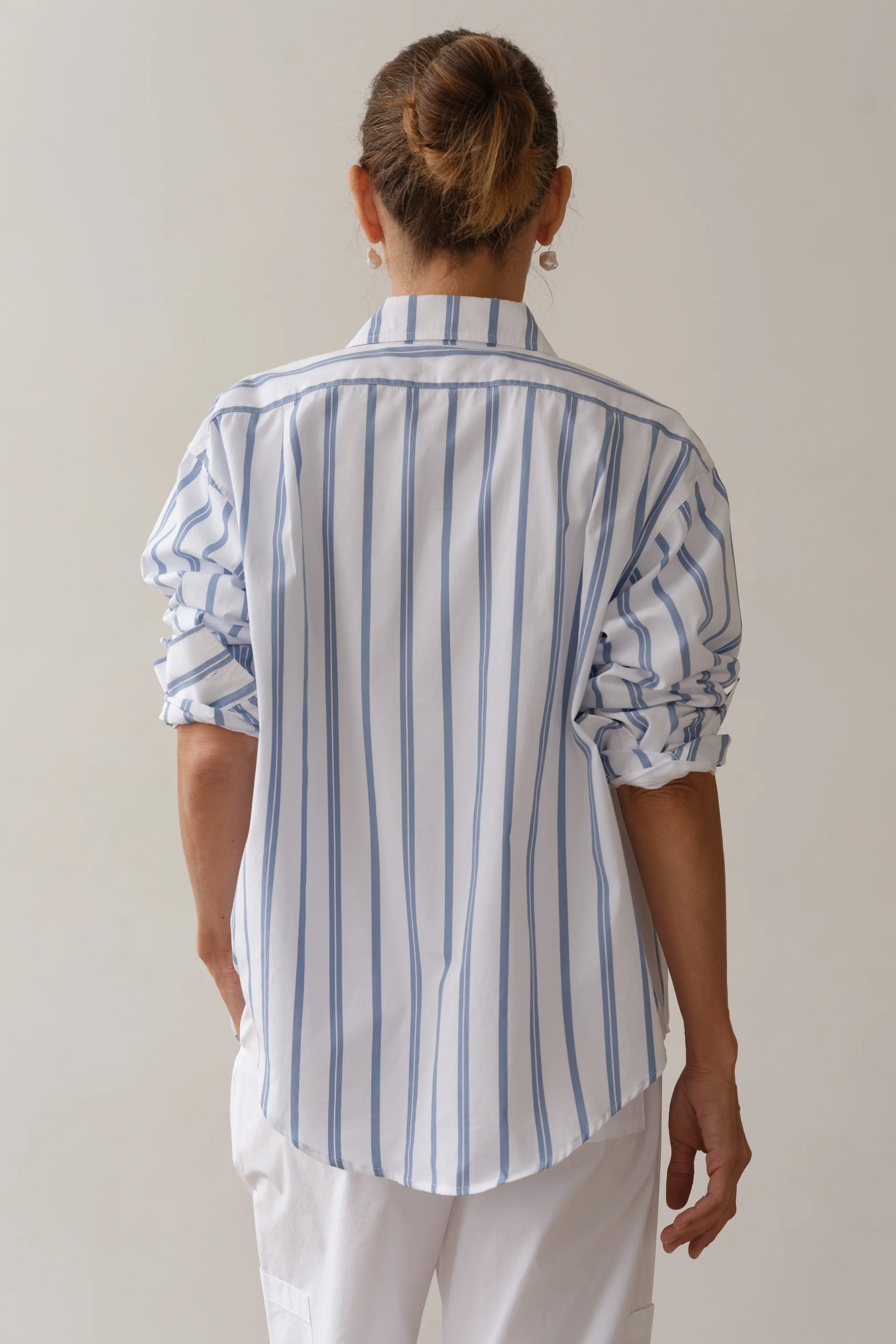 A person with hair in a bun wears Donni's The Poplin Shirt, featuring blue and white vertical stripes and crafted from cotton poplin for a vintage men's shirting vibe. With rolled-up sleeves and a relaxed fit, it's paired effortlessly with white pants against a plain light background.