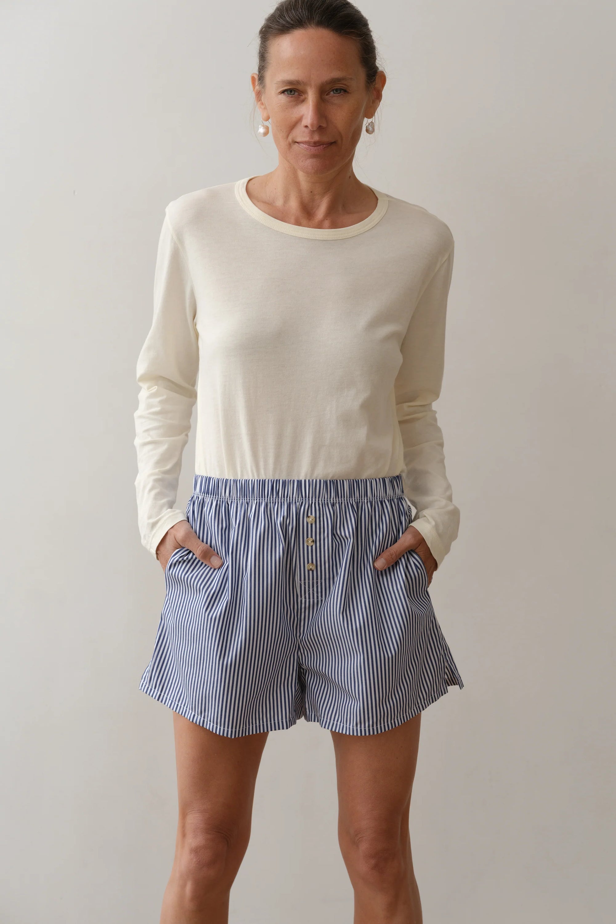 In front of a plain backdrop, a person wears the cream long-sleeve top and Donni's The Stripe Pop Boxer. Made of lightweight breathable cotton poplin, their outfit pairs well with their short hair and hoop earrings as they strike a neutral pose with hands in pockets.