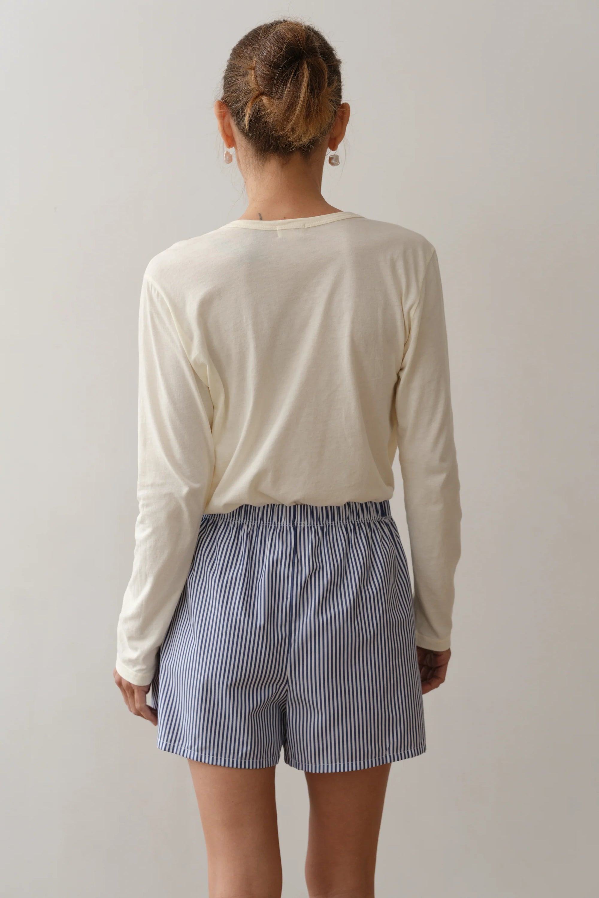 A person stands facing away wearing a lightweight, breathable cream-colored cotton poplin shirt and "The Stripe Pop Boxer" blue-and-white striped shorts by Donni. The relaxed fit complements the neutral background, and their hair is tied in a neat bun.