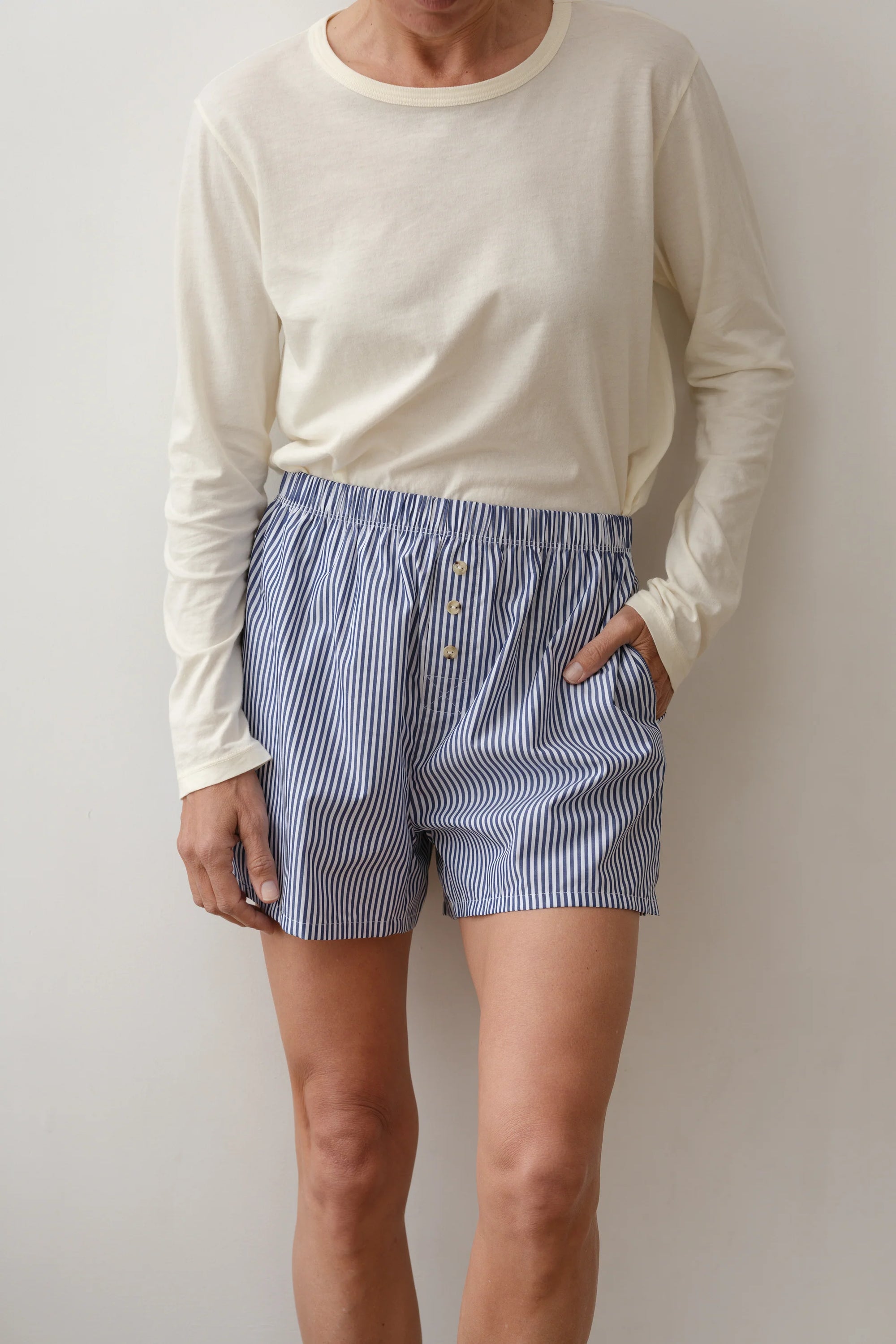 A person in an off-white cotton poplin long-sleeve shirt and Donni's The Stripe Pop Boxer, featuring a relaxed fit in blue-and-white stripes, stands against a plain wall. One hand is in a pocket; the other hangs by their side. The image is cropped at the shoulders.