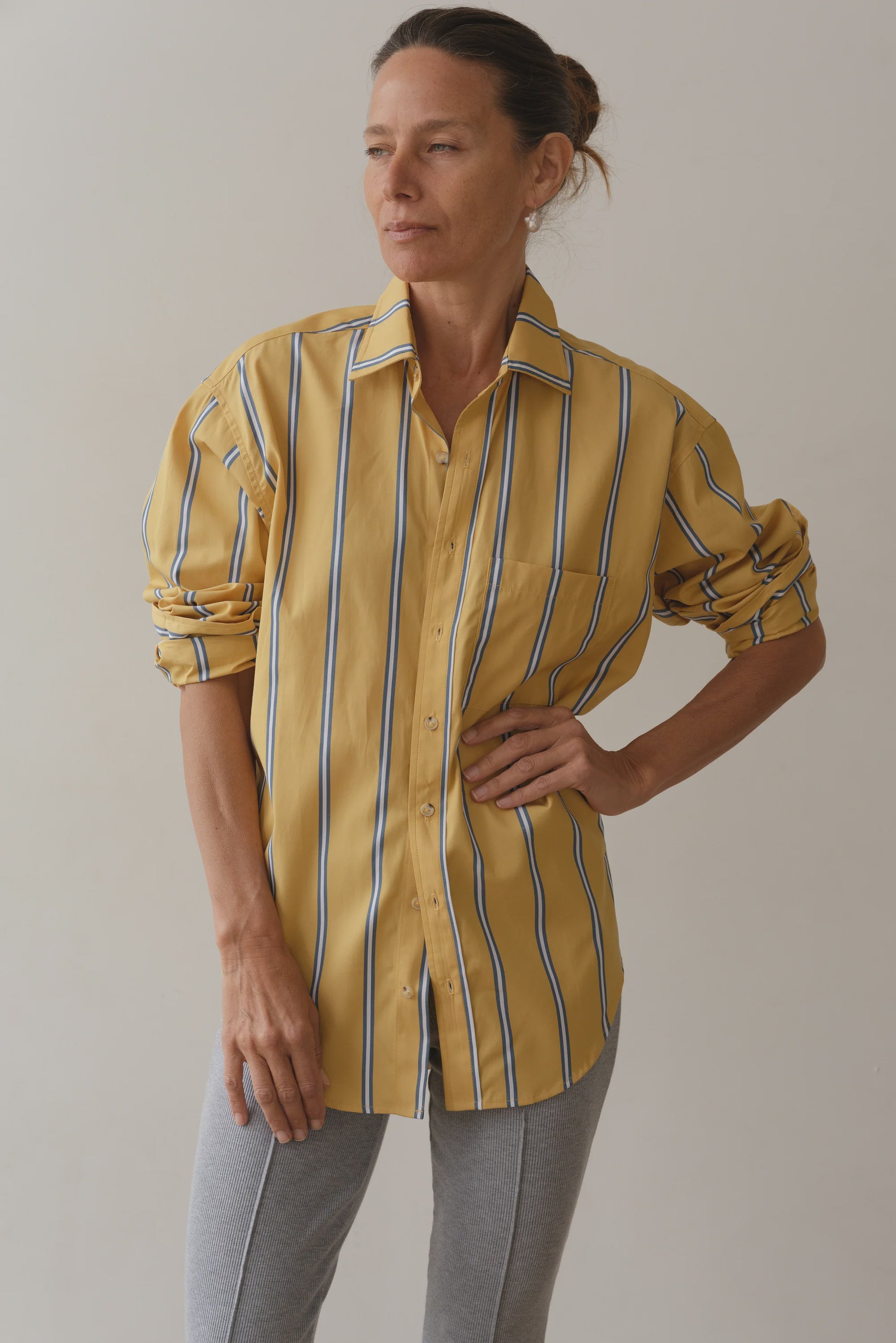 The individual dons The Poplin Shirt by Donni—a relaxed fit, yellow striped vintage men's shirting style with rolled-up sleeves—and pairs it with gray pants. They stand against a plain background, looking to the side.