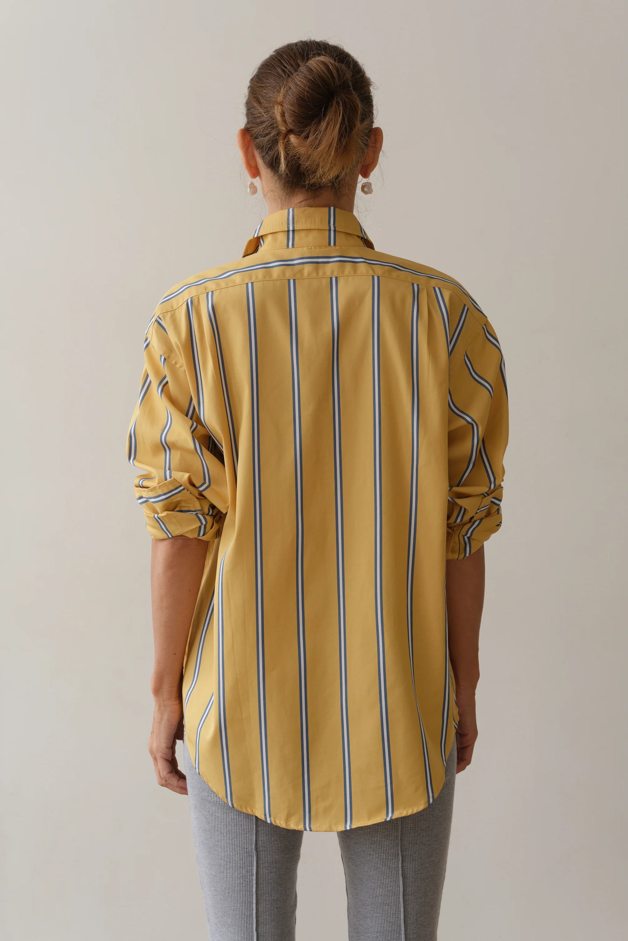 A person with their hair in a bun is seen from the back wearing Donni's The Poplin Shirt, featuring yellow, blue, and white vertical stripes of vintage men's shirting fabric. Paired with gray pants against a plain backdrop, they display a relaxed and effortlessly stylish fit.