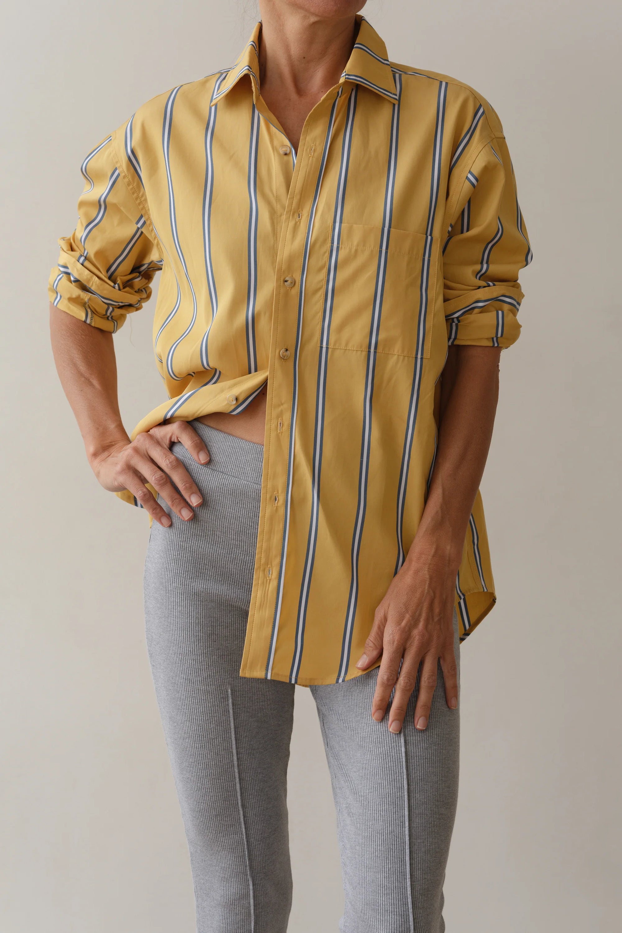 A person in Donni's The Poplin Shirt, featuring a yellow and blue stripe pattern on cotton poplin, pairs it with light gray pants. The oversized shirt has a relaxed fit, rolled sleeves, and is partially tucked, with one hand on their hip.