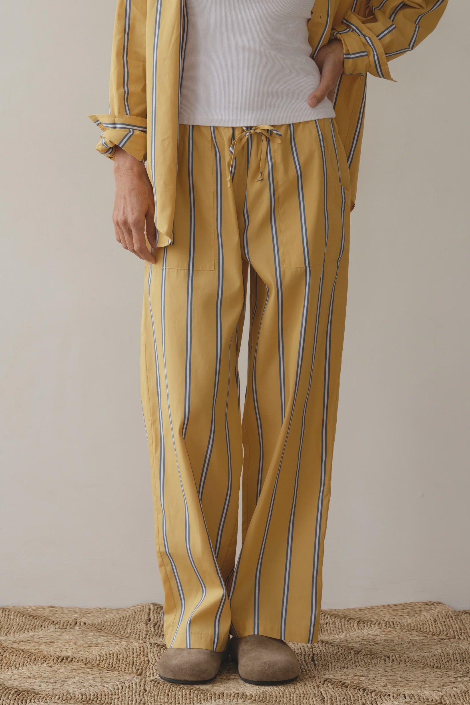 The person, showcasing a versatile style for relaxed comfort, is dressed in loose-fitting mustard yellow "The Stripe Pop Drawstring Pant" by Donni, made of cotton poplin. They pair it with a matching top over a white shirt, stand on a woven rug, and wear brown slippers.