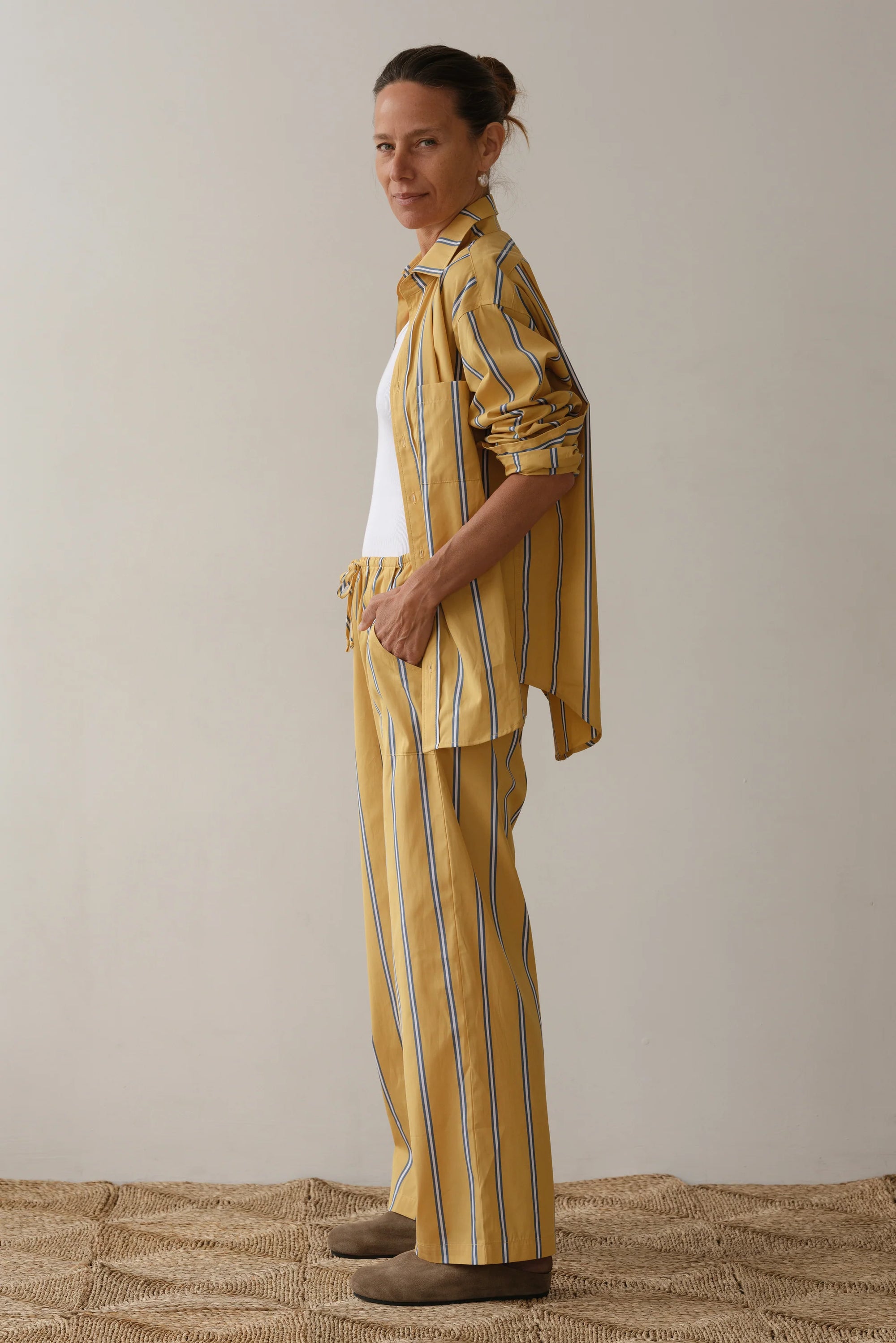 A relaxed individual with tied-up brown hair wears a yellow striped shirt and Donni's The Stripe Pop Drawstring Pant made of cotton poplin over a white top. They stand barefoot on a textured surface against a plain, light background.