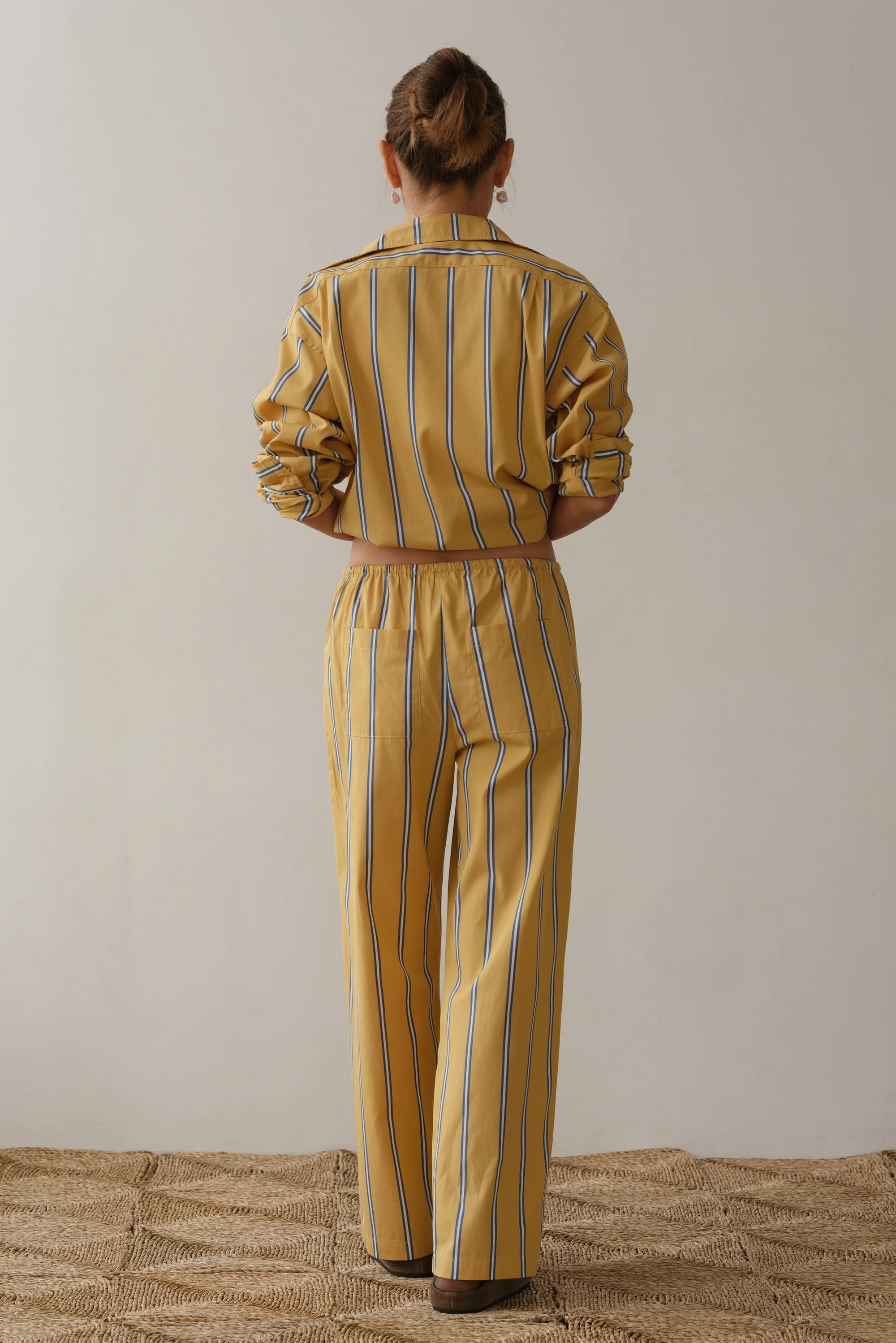 A person with hair in a bun, seen from behind, wears a yellow and white striped outfit by Donni, featuring soft cotton poplin with long sleeves and The Stripe Pop Drawstring Pant. The pants have a comfy drawstring waist that enhances the woven texture against the plain backdrop.