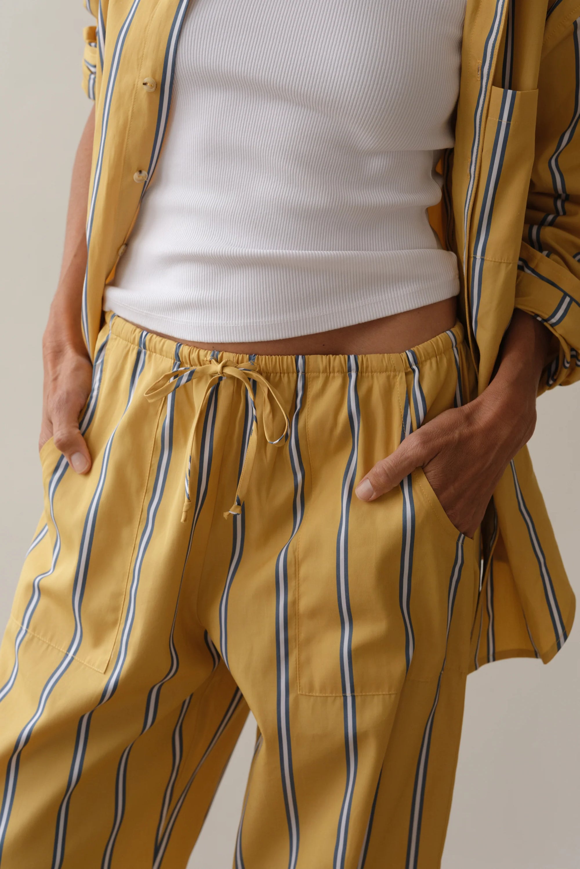 Donni's The Stripe Pop Drawstring Pant, in soft cotton poplin with blue and white stripes, offers comfort with its drawstring waist. Paired with a partially unbuttoned white ribbed tank top, this relaxed yellow pajama set allows easy hand-tucking into pockets.