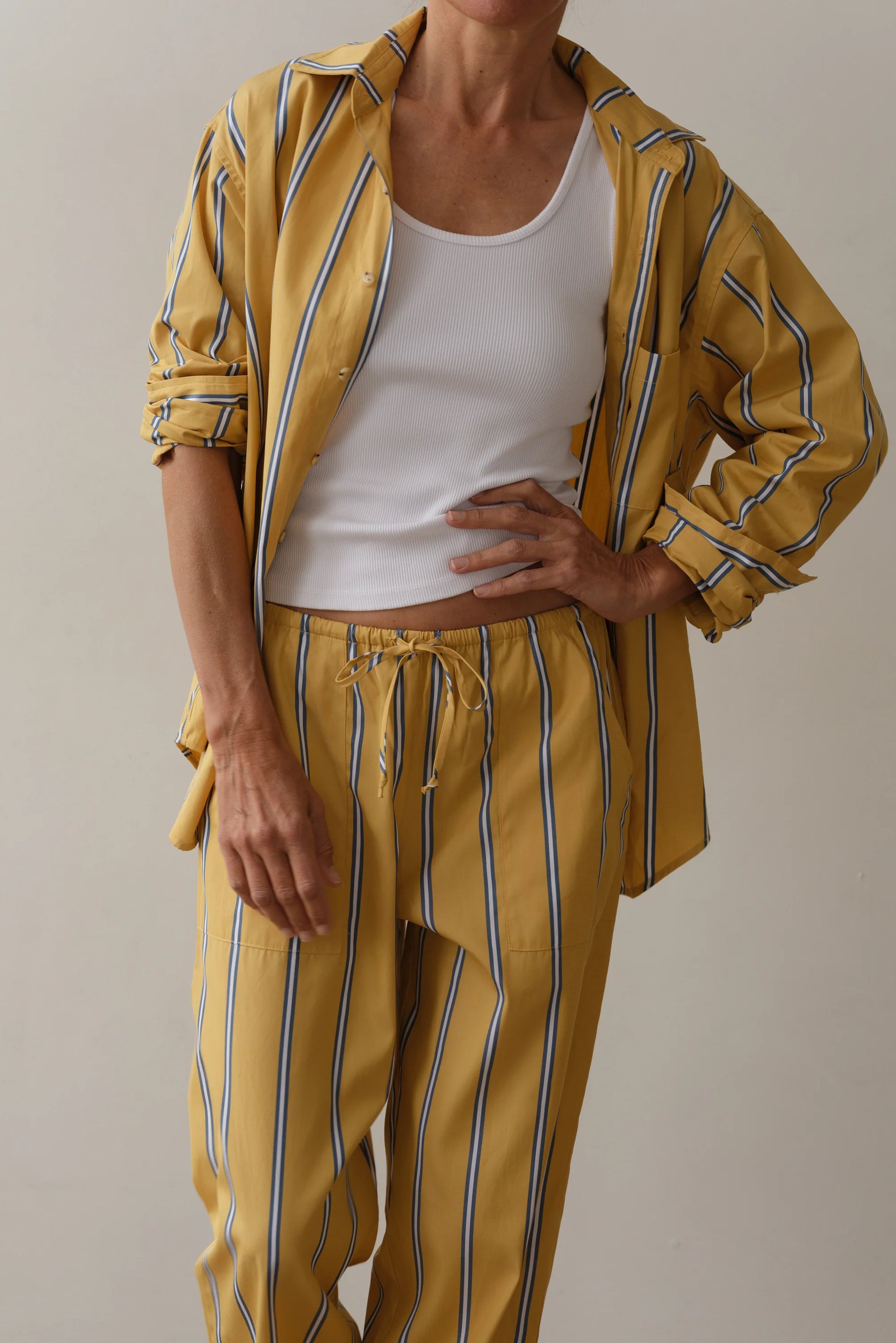 A person wears Donni's The Stripe Pop Drawstring Pant, featuring a loose yellow design with blue and white vertical stripes. Paired with a white tank top, their left hand is on their hip while the right is relaxed by their side, exuding casual elegance in cotton poplin.