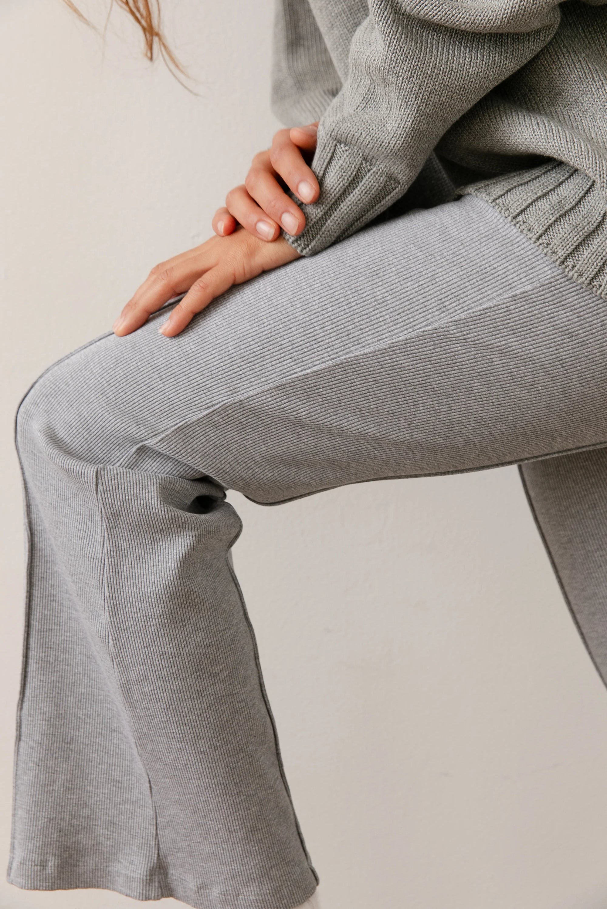 A person in Donni&#39;s The Rib Kick Flare set, featuring a gray ribbed sweater and matching high-rise pants, sits with their hand resting gently on their knee. The soft rib fabric looks perfect for cozy or casual vibes.