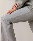 A person in Donni's The Rib Kick Flare set, featuring a gray ribbed sweater and matching high-rise pants, sits with their hand resting gently on their knee. The soft rib fabric looks perfect for cozy or casual vibes.