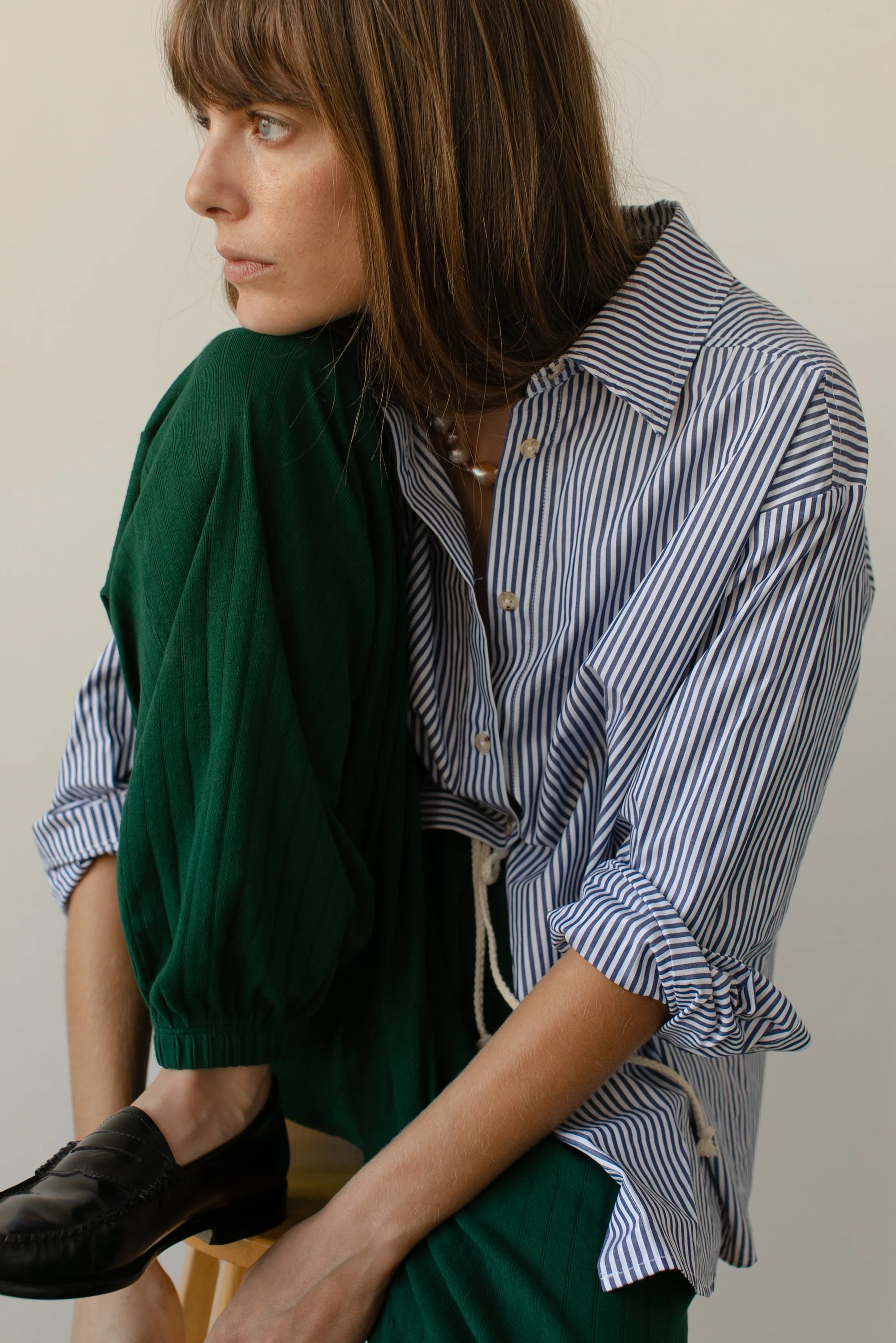 A person with long brown hair sits sideways, thoughtfully gazing into the distance. They&#39;re wearing The Stripe Pop Shirt by Donni, featuring tortoise-like buttons, and green pants, with one leg resting on a wooden surface indoors against a neutral background.