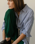 A person with long brown hair sits sideways, thoughtfully gazing into the distance. They're wearing The Stripe Pop Shirt by Donni, featuring tortoise-like buttons, and green pants, with one leg resting on a wooden surface indoors against a neutral background.