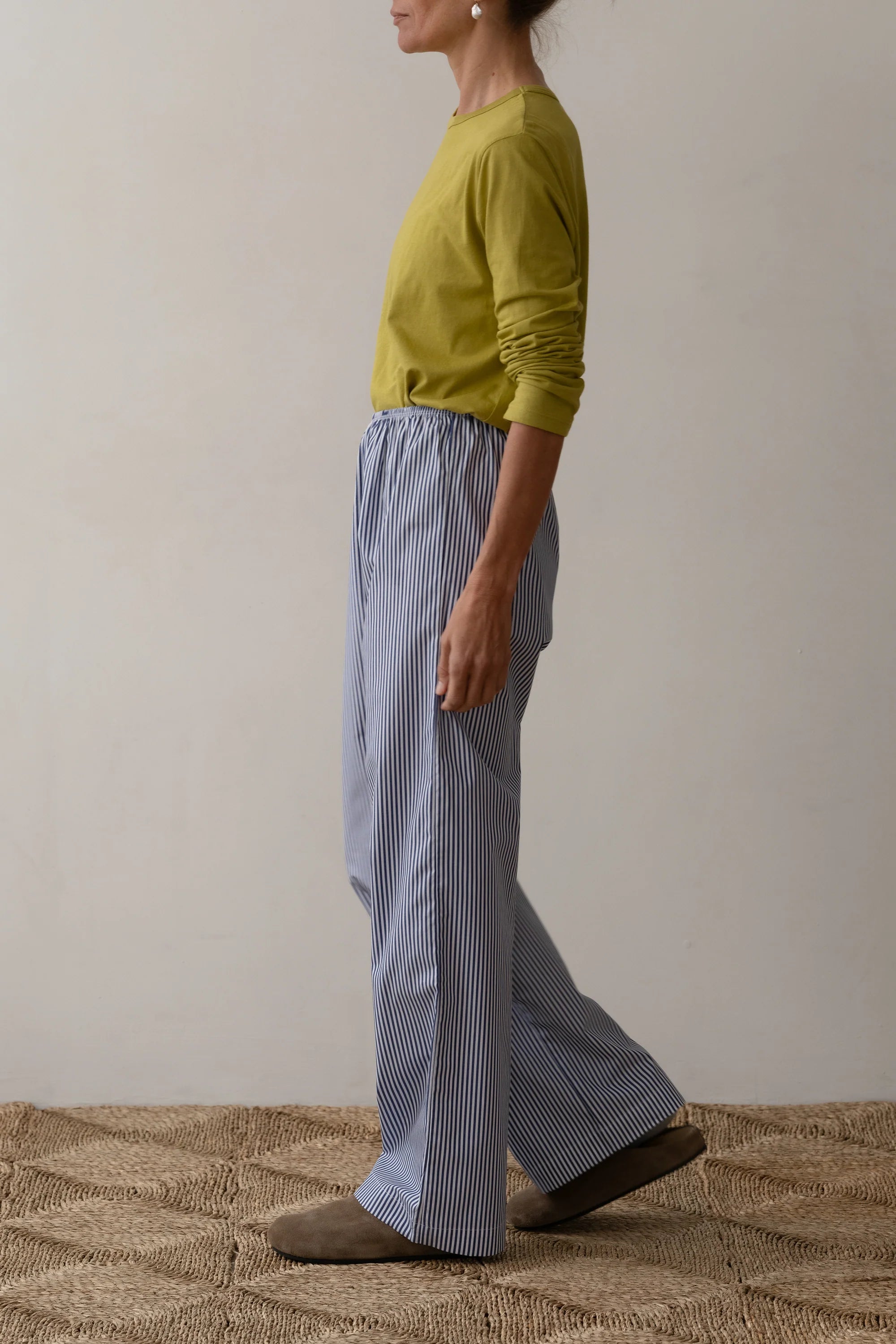 A woman in a yellow long-sleeve shirt, Donni Stripe Pop Pant XS, and brown shoes stands sideways on a woven rug against a plain light-colored wall.