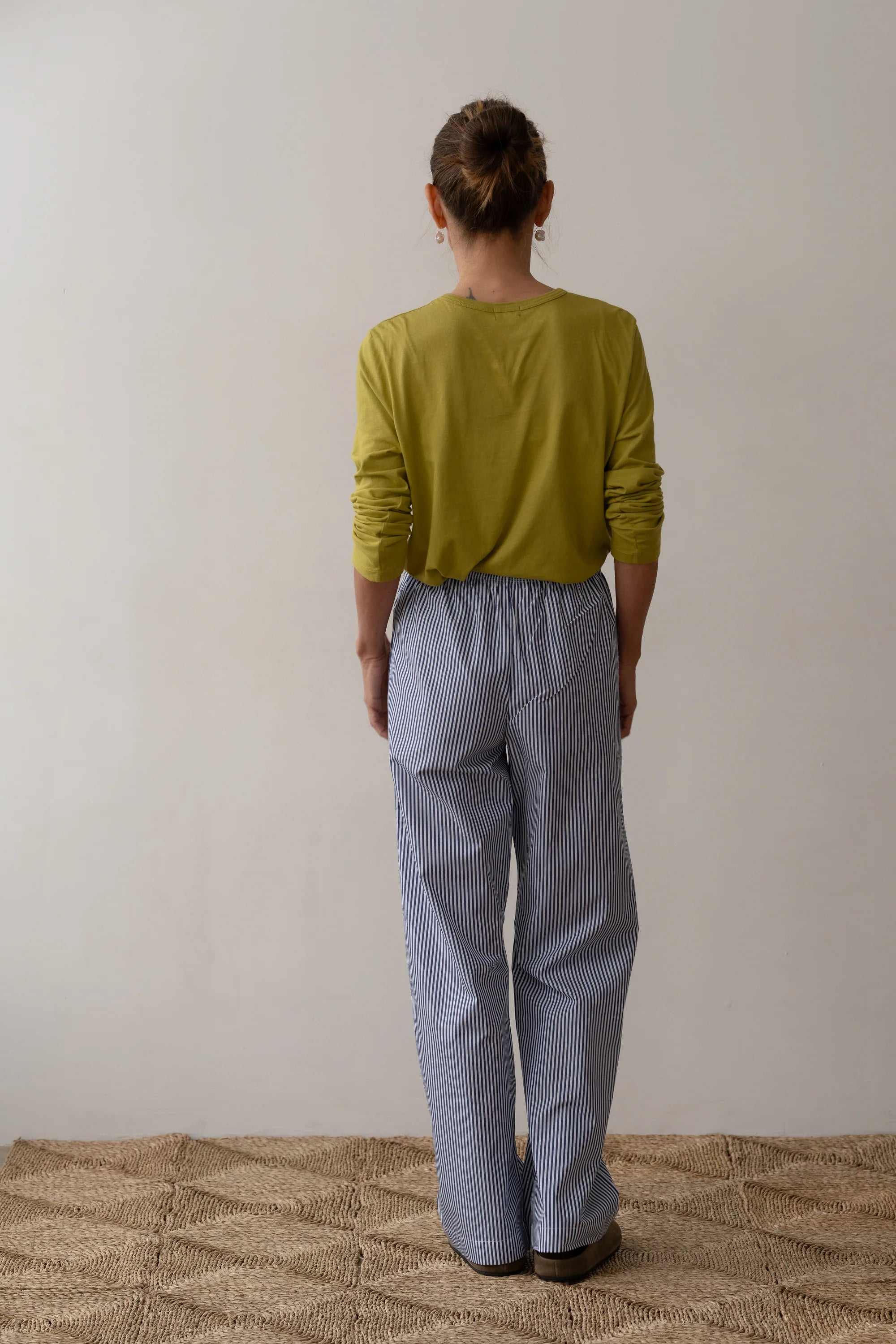 A person with brown hair in a bun wears the Donni Stripe Pop Pant XS, featuring blue-and-white stripes and an elastic waistband, paired with a long-sleeved green shirt while standing barefoot on a woven rug facing a light wall.