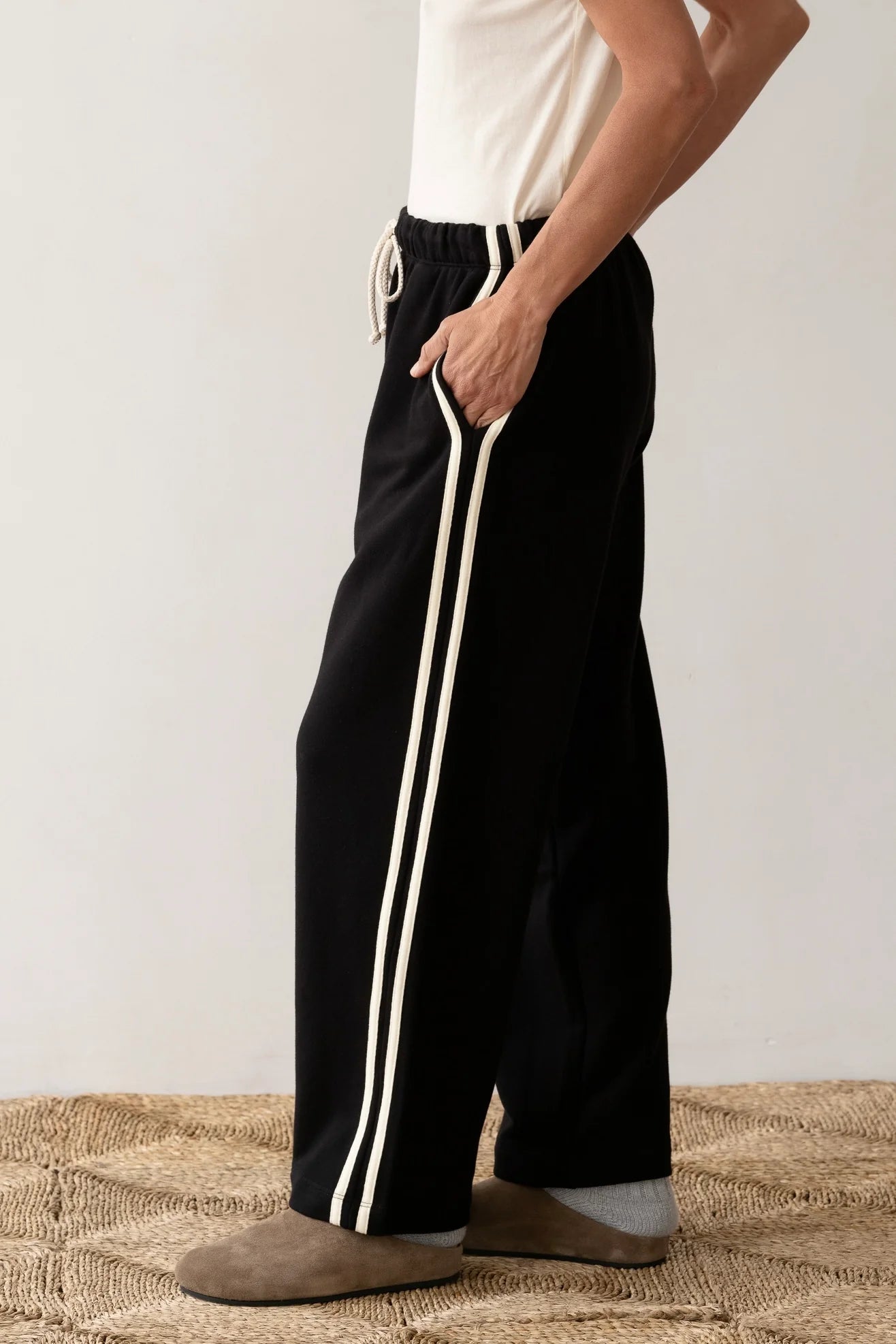 Donning Donni's The Eco Terry Stripe Crop Pant featuring white side stripes and a white organic cotton T-shirt, a person stands with one hand in their pocket. They sport tan slip-on shoes against a plain backdrop with a woven rug underfoot.