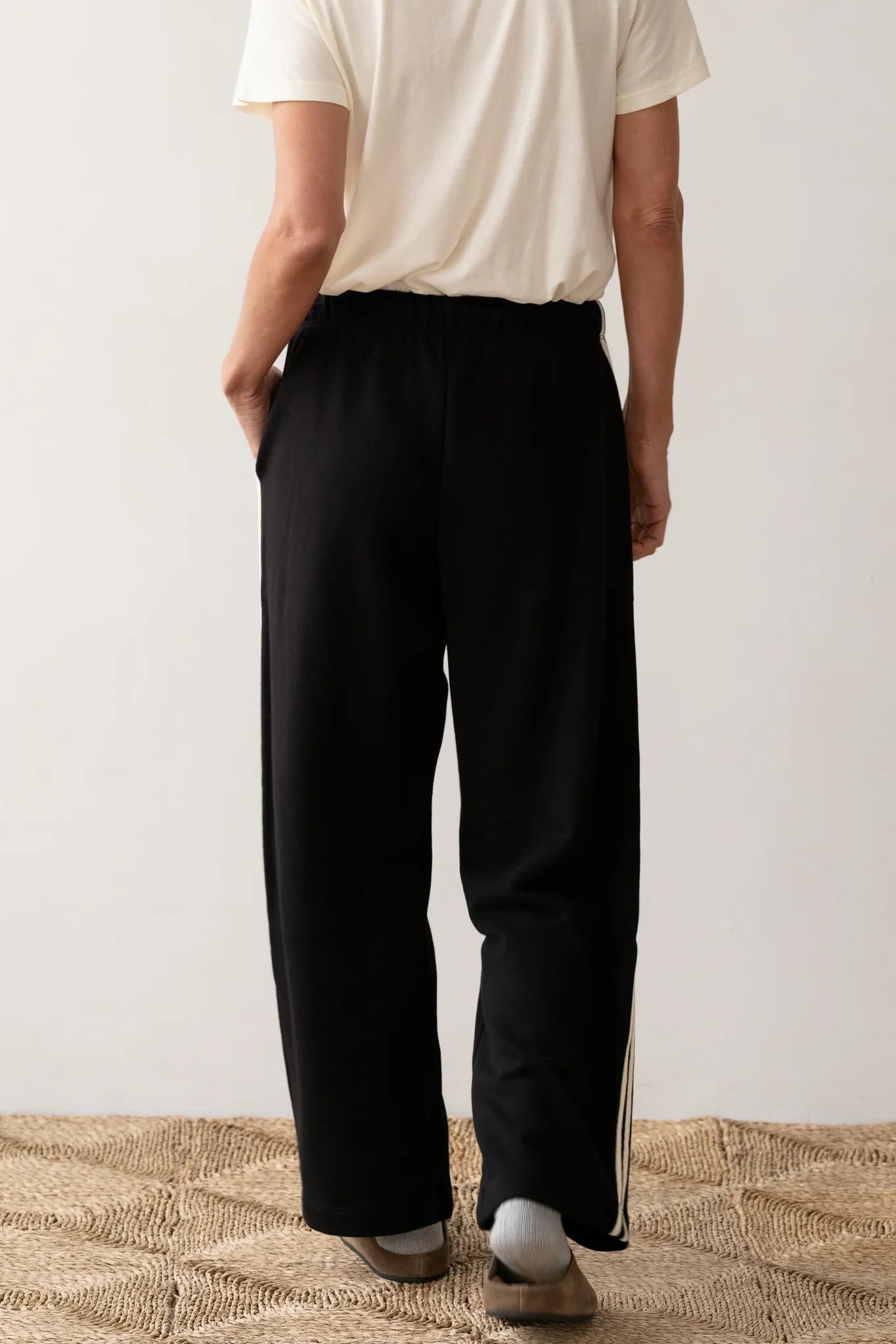 Standing on a woven rug against a plain white backdrop, a person wearing Donni's white T-shirt and The Eco Terry Stripe Crop Pant poses with hands in pockets. The loose black pants, crafted from organic cotton, radiate an air of effortless style and comfort.