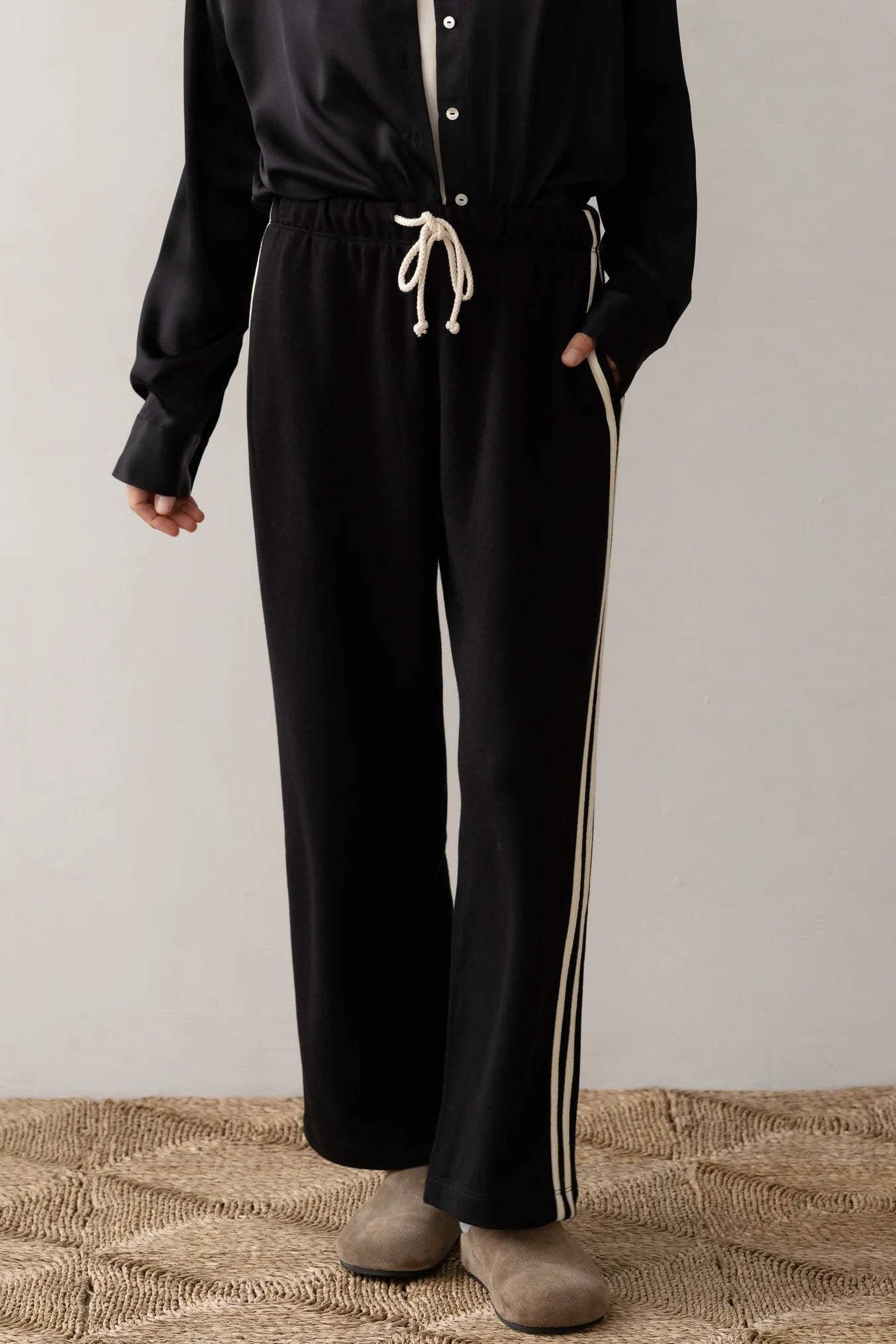 A person is wearing Donni's The Eco Terry Stripe Crop Pant, a black wide-leg crop pant with white stripes featuring a drawstring waist, paired with a black button-up shirt. They stand on a textured carpeted floor, wearing beige shoes against a plain background.