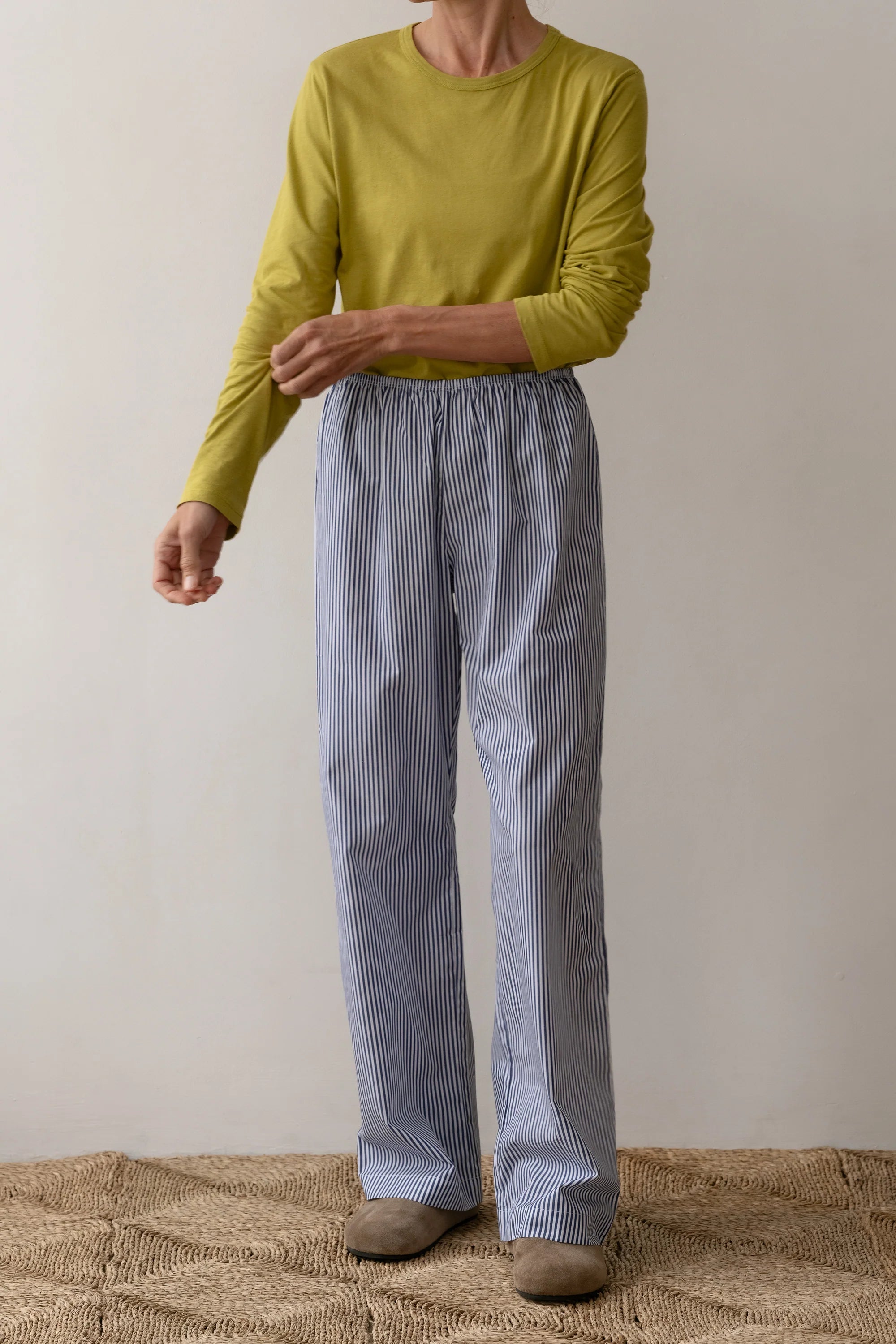 A person stands on a woven rug against a plain background, wearing the Donni Stripe Pop Pant XS—loose, high-waisted blue-and-white striped cotton poplin pants with a straight-leg fit. Their face is not visible; one hand adjusts their sleeve.