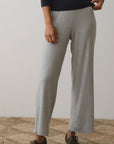 A person dressed in The Baby Rib Scallop Pant by Donni, featuring a black long-sleeve top with a cropped fit and gray wide-leg pants, stands on a textured beige rug. They wear dark brown sandals that contrast against the plain, light-colored background.