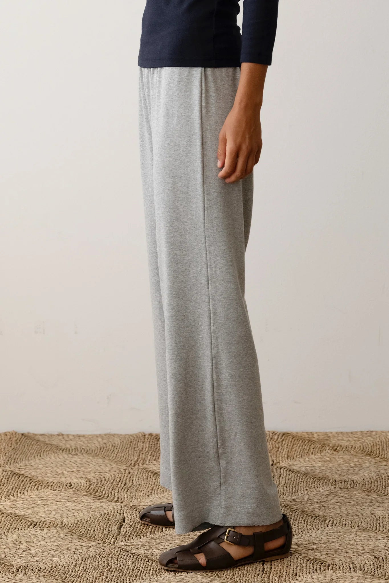 A person stands on a textured floor, clad in black long-sleeve shirt and The Baby Rib Scallop Pant by Donni in gray, along with brown sandals. The image is cropped to display only the lower half of the body.