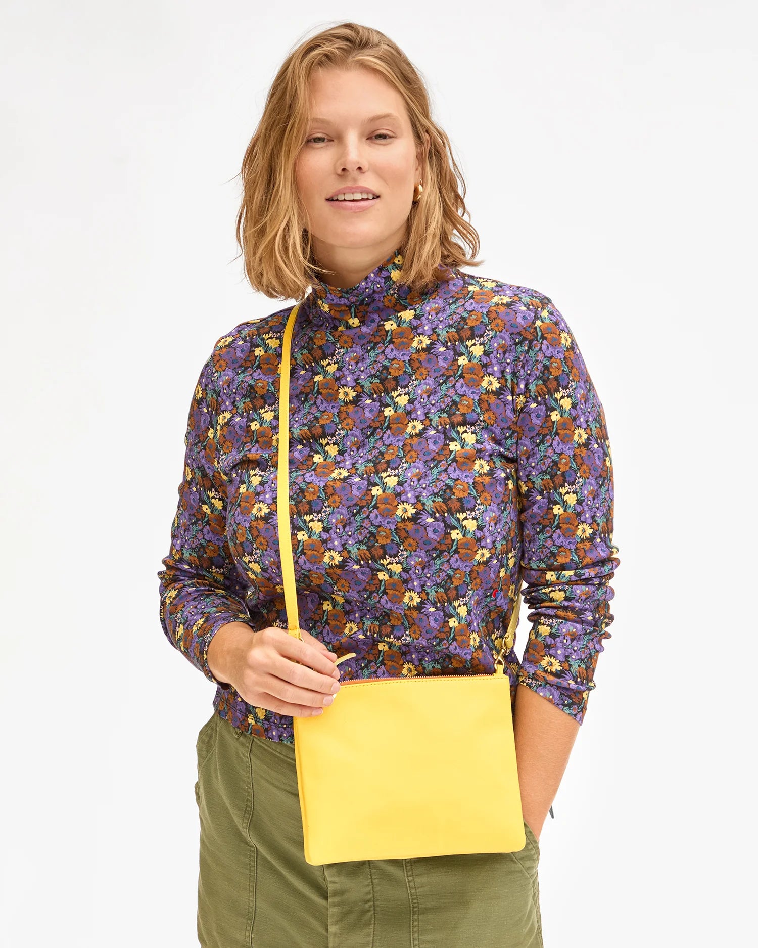 A person with shoulder-length hair, adorned in a vibrant floral long-sleeve top and green pants, is holding the Clare Vivier Double Sac Bretelle crossbody bag with a detachable shoulder strap. They are smiling and facing the camera against a plain white background.