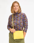 A person with shoulder-length hair, adorned in a vibrant floral long-sleeve top and green pants, is holding the Clare Vivier Double Sac Bretelle crossbody bag with a detachable shoulder strap. They are smiling and facing the camera against a plain white background.