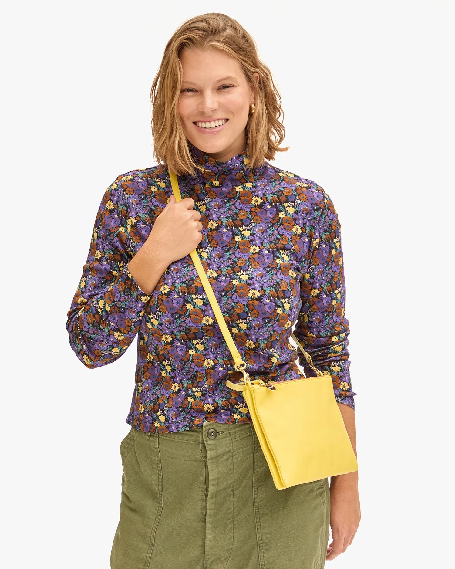 A person with shoulder-length blonde hair smiles while wearing a long-sleeved, purple floral top, green pants, and the Clare Vivier Double Sac Bretelle yellow crossbody bag with a detachable shoulder strap. The background is plain white.