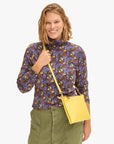 A person with shoulder-length blonde hair smiles while wearing a long-sleeved, purple floral top, green pants, and the Clare Vivier Double Sac Bretelle yellow crossbody bag with a detachable shoulder strap. The background is plain white.