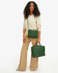 A woman with curly hair stands against a white background wearing a beige sweater, tan flared pants, and leopard print shoes. She carries the elegant Clare Vivier Double Sac Bretelle handbag made from Italian leather with double zippered pouches, along with a small green crossbody bag.