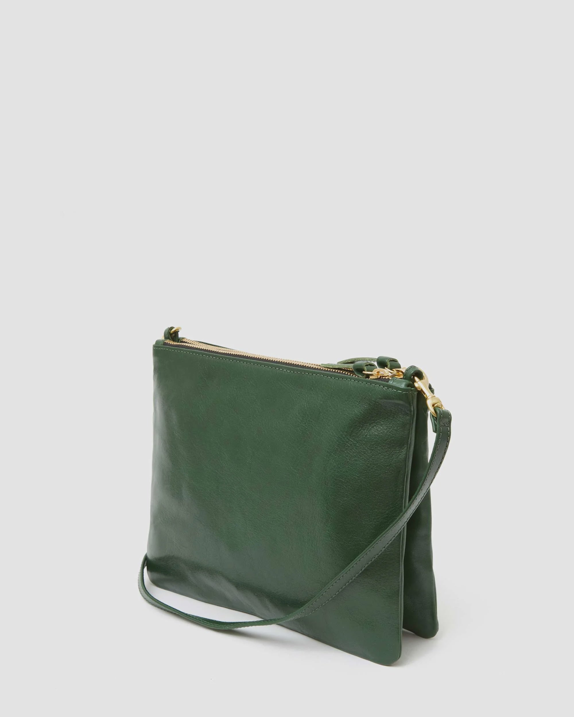 The Double Sac Bretelle by Clare Vivier is beautifully showcased against a plain light grey background. Its sleek, dark green Italian leather exterior boasts a minimalist design with a single strap and a gold zipper closure. The bag is slightly folded to reveal its elegant side profile.