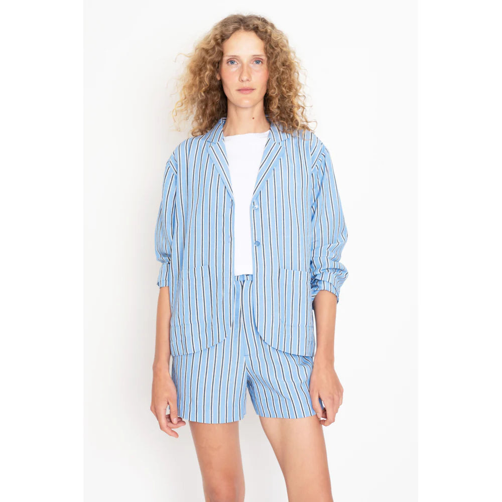 A person with curly hair wears Kule's The Gwen blazer in royal blue, beige, and navy stripes with matching trouser shorts over a white top, set against a plain white background.