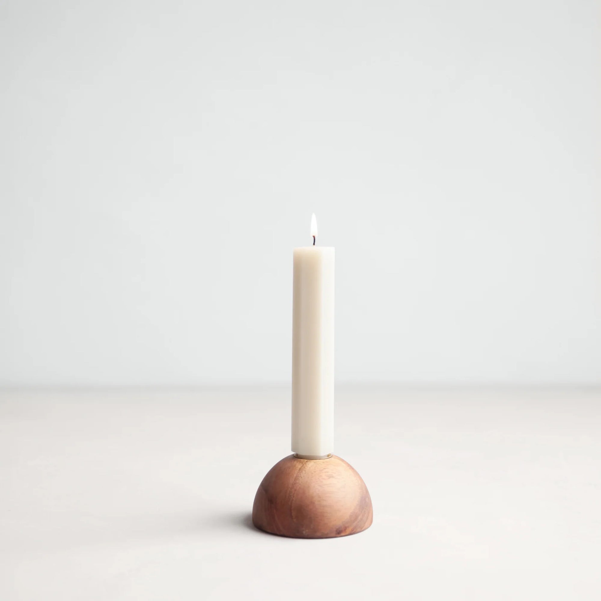 A single white candle is lit and placed in The Floral Society's Dusk Taper Holder, a hand-finished walnut piece, set against a plain light gray background.