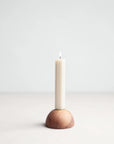 A single white candle is lit and placed in The Floral Society's Dusk Taper Holder, a hand-finished walnut piece, set against a plain light gray background.