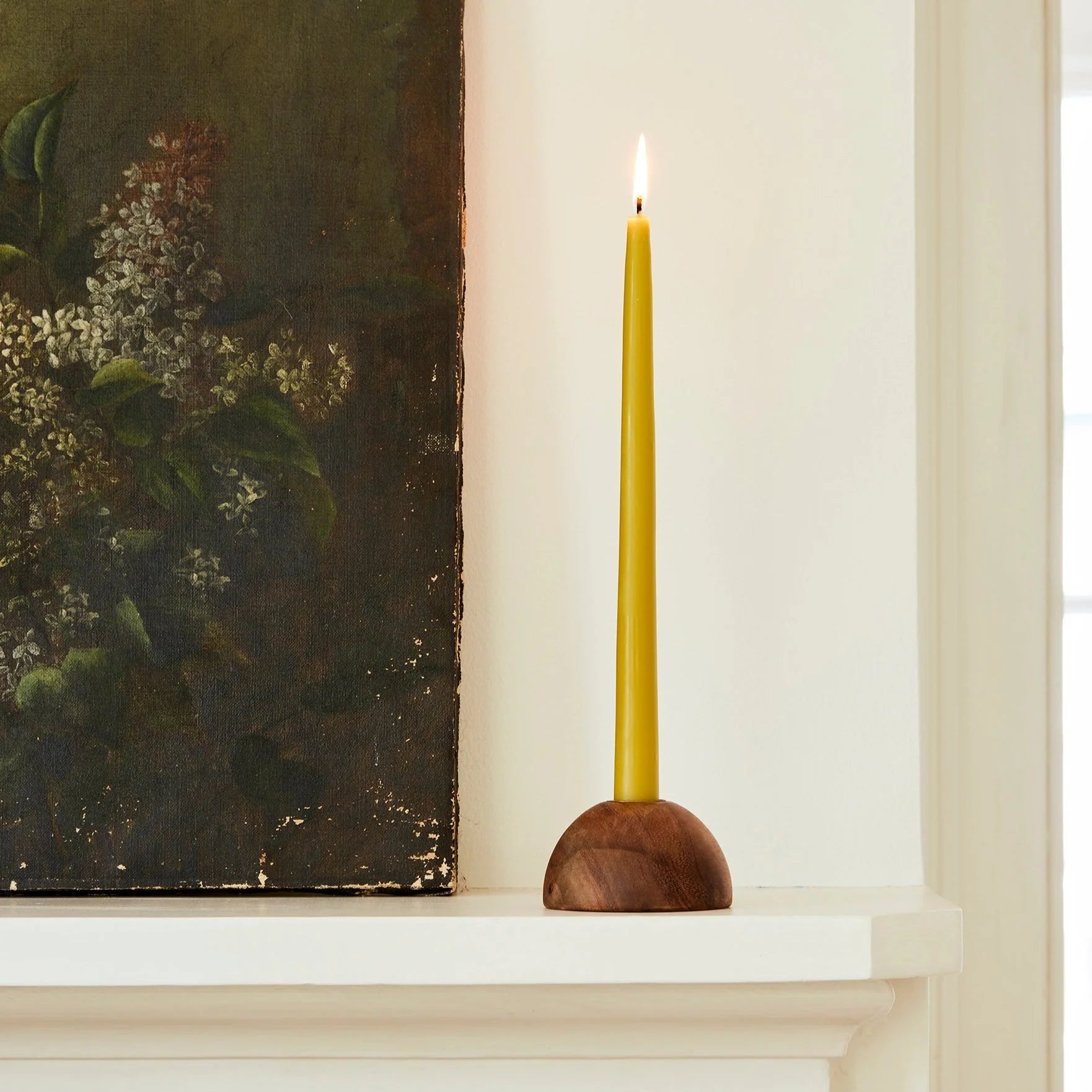A tall yellow candle in The Floral Society’s Dusk Taper Holder sits lit on a white mantle beside a vintage-style floral painting with a dark background, as light pours through the window behind.