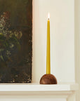 A tall yellow candle in The Floral Society’s Dusk Taper Holder sits lit on a white mantle beside a vintage-style floral painting with a dark background, as light pours through the window behind.