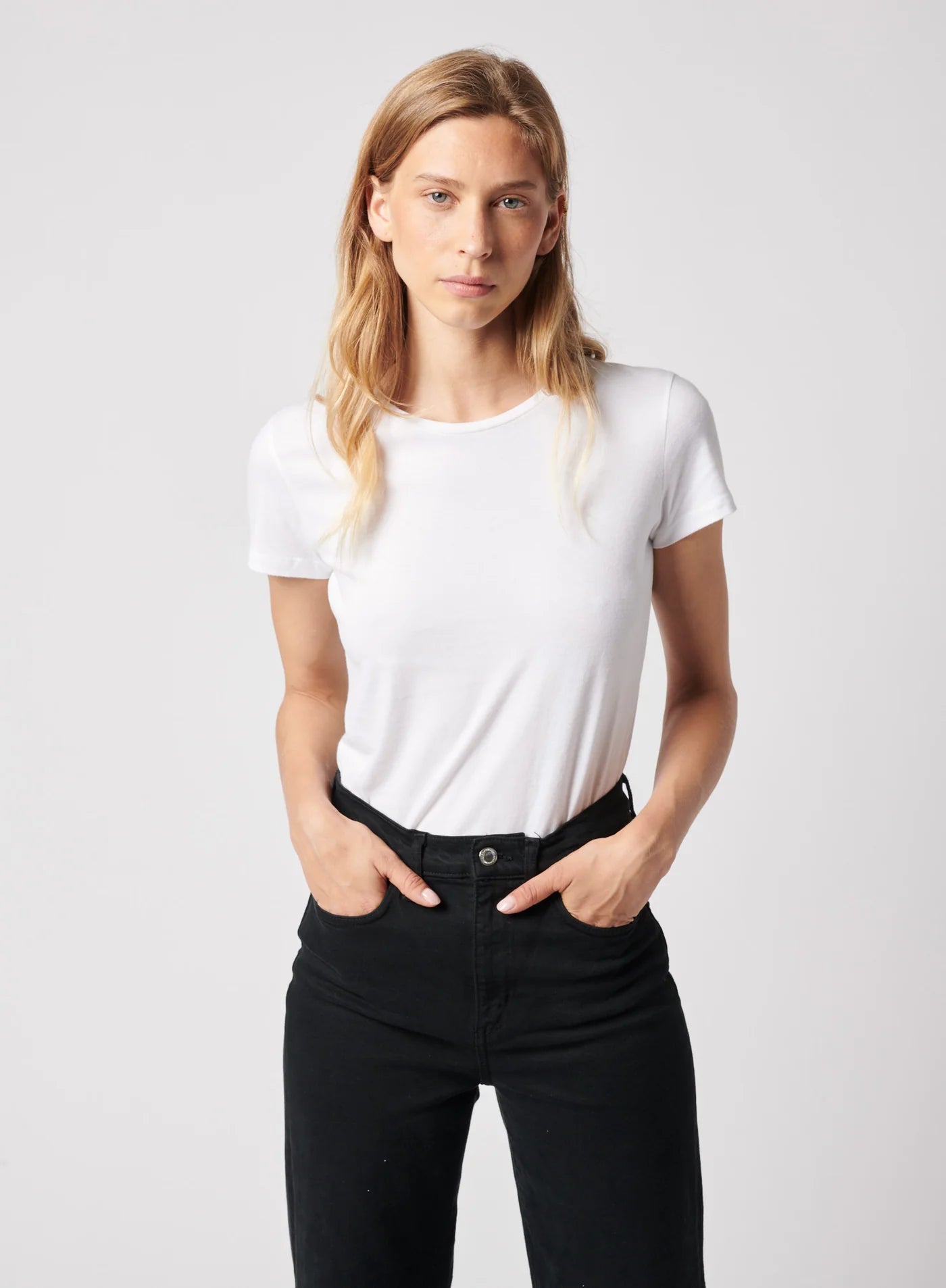 A person with long blonde hair wears the Majestic Filatures Deluxe Cotton S/S Crewneck and high-waisted black pants, standing against a light grey background with hands in pockets and looking at the camera.