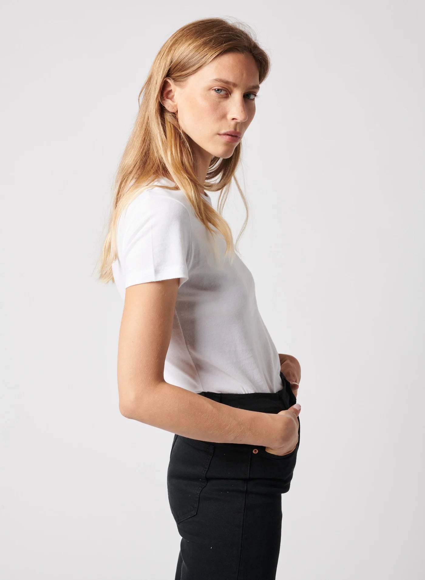 A woman with long blonde hair wears a white Majestic Filatures Deluxe Cotton S/S Crewneck tee and black pants, standing in profile against a plain light background, looking seriously at the camera.