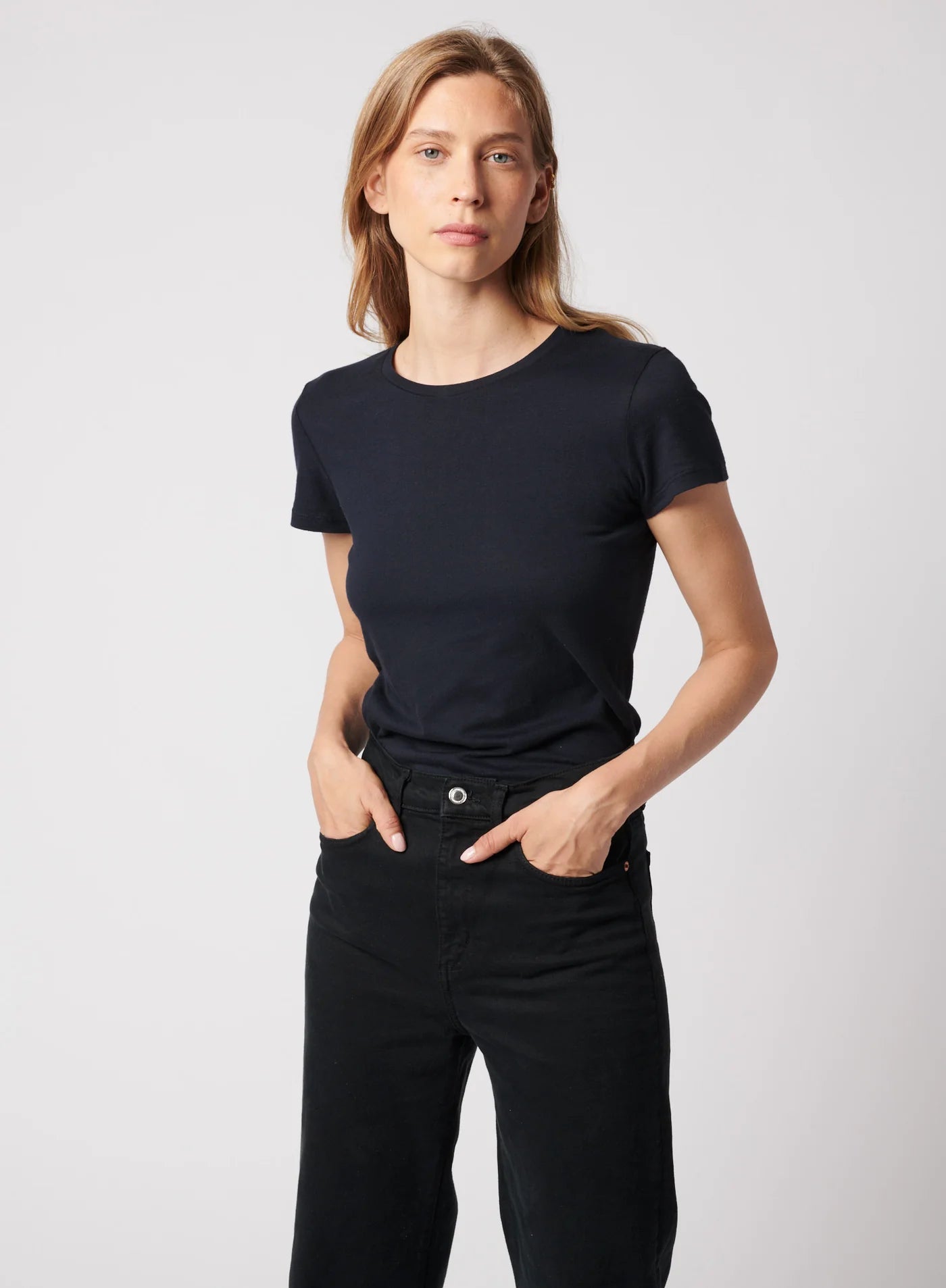 A person with long, light brown hair stands against a plain background, wearing Majestic Filatures’ Deluxe Cotton S/S Crewneck in black and black pants, hands in pockets, looking confidently at the camera.