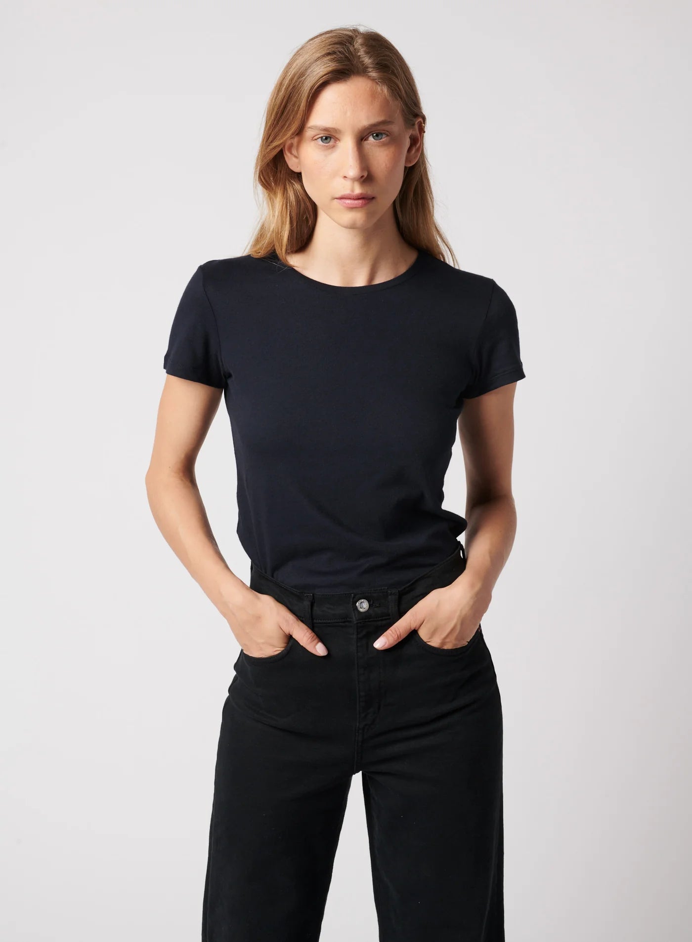 A person with long blonde hair wears the Majestic Filatures Deluxe Cotton S/S Crewneck in black, paired with black pants, standing hands-in-pockets and facing the camera against a plain light background.