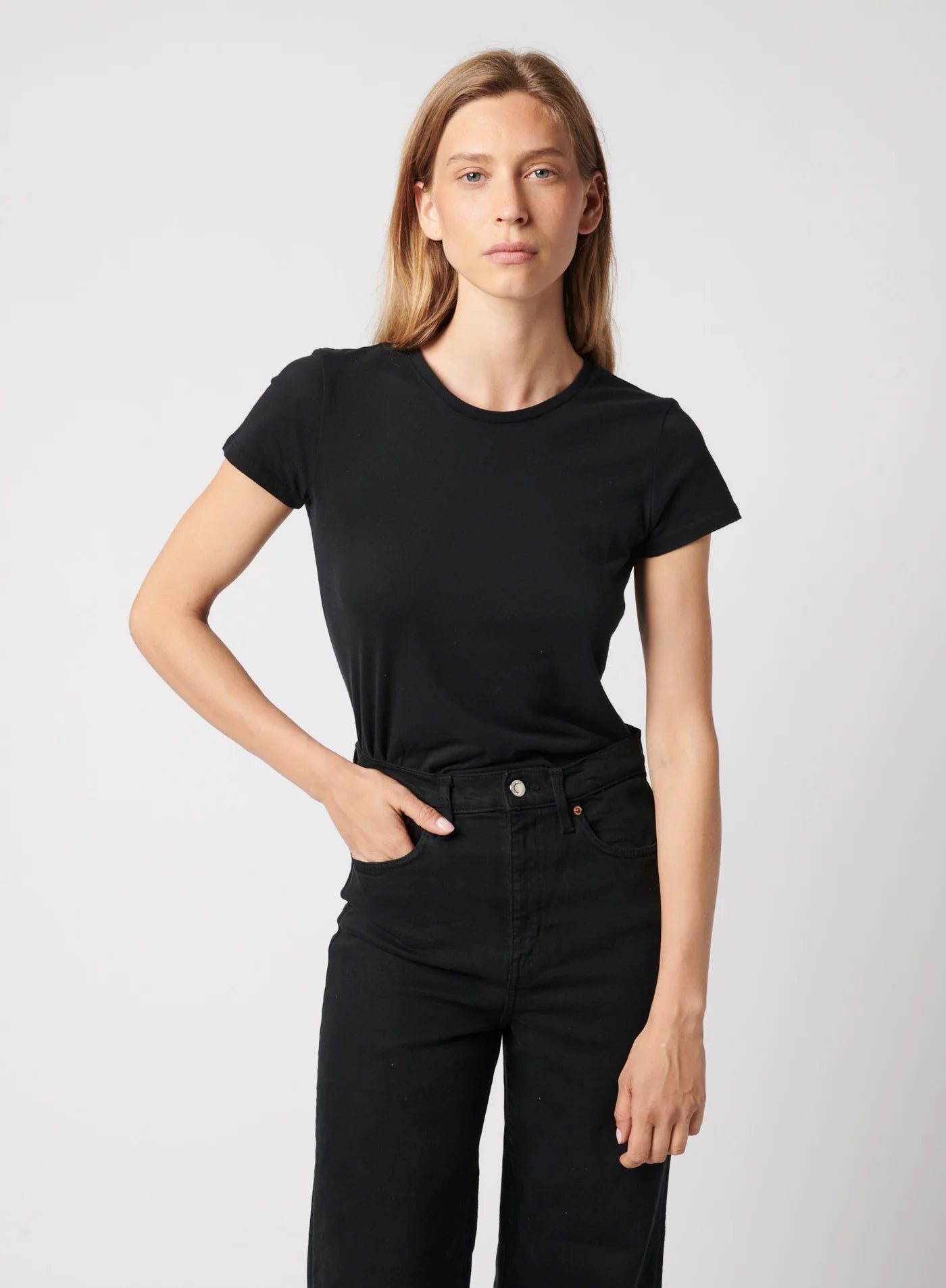 A person with long light brown hair wears the Majestic Filatures Deluxe Cotton S/S Crewneck and black jeans, standing against a plain light background, looking directly at the camera with a neutral expression and one hand in their pocket.