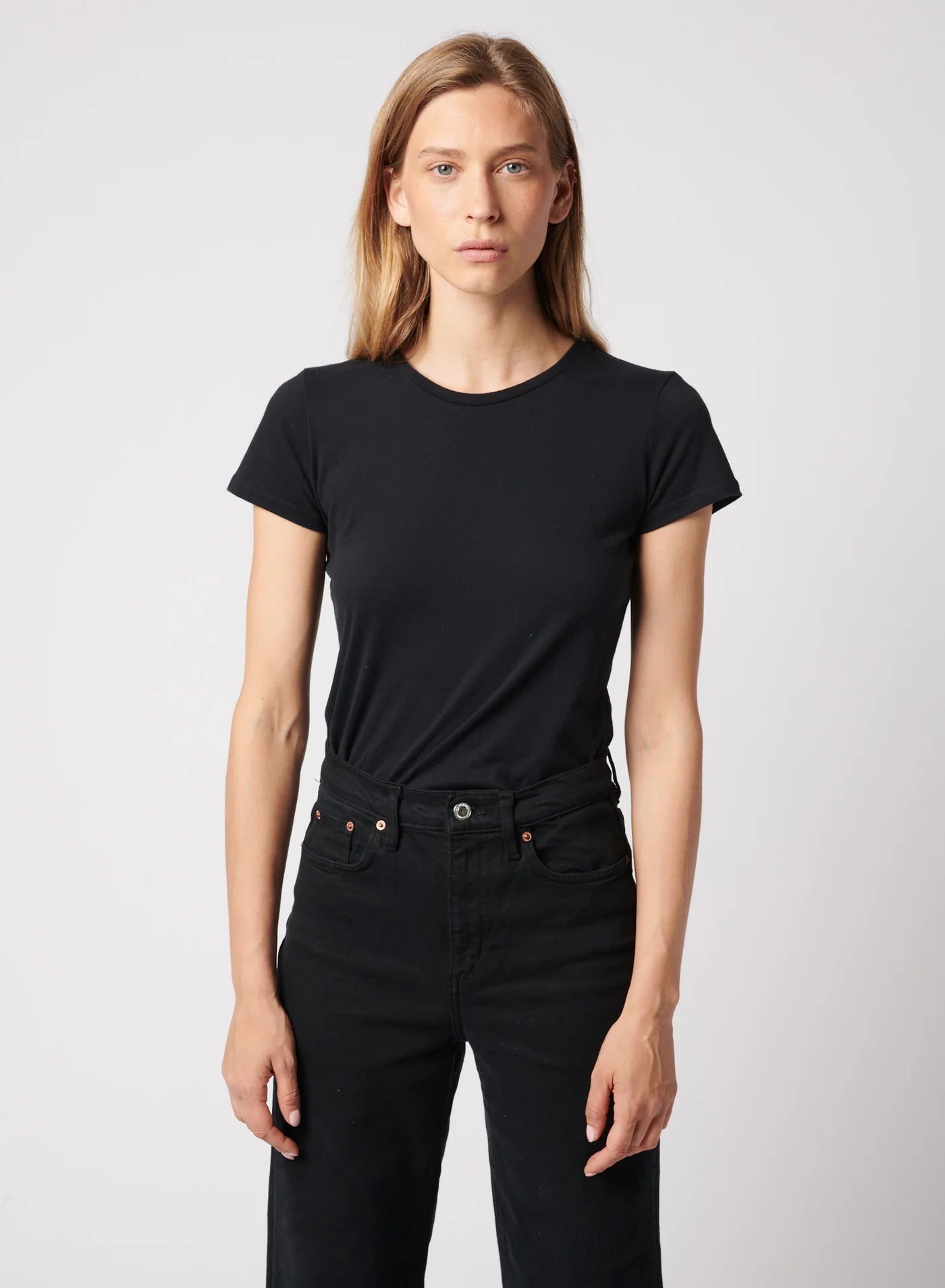 A person with long, straight blonde hair wears the Majestic Filatures Deluxe Cotton S/S Crewneck in black and black pants, standing against a light gray background and looking directly at the camera with a neutral expression.