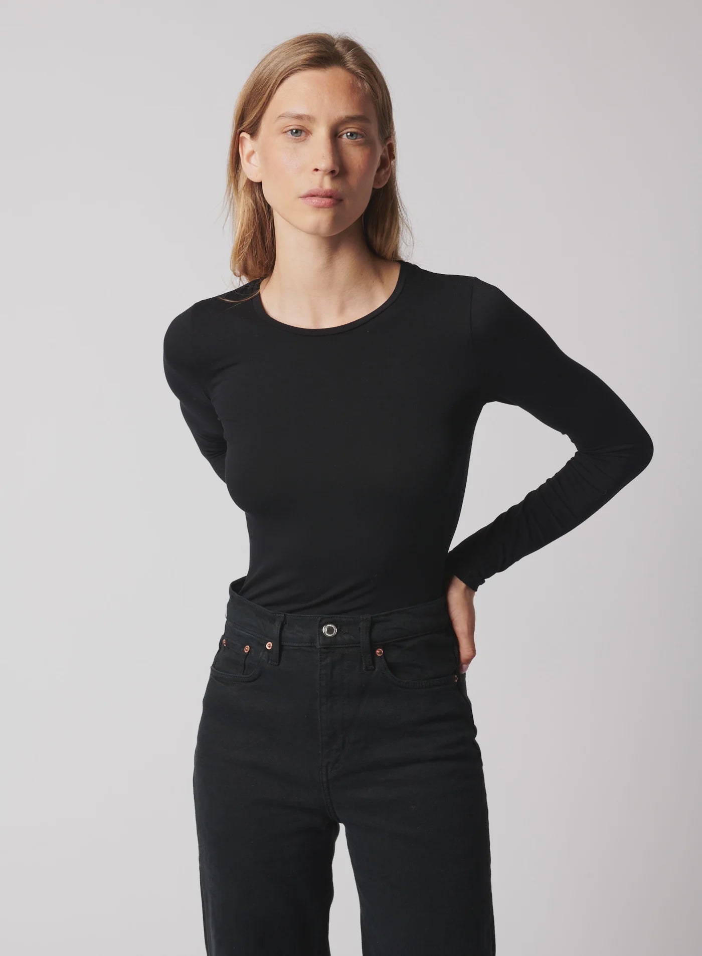 A person with straight light brown hair wears the Majestic Filatures Soft Touch L/S Crewneck T-Shirt in black and black jeans, standing against a plain light gray background and looking at the camera with a neutral expression.